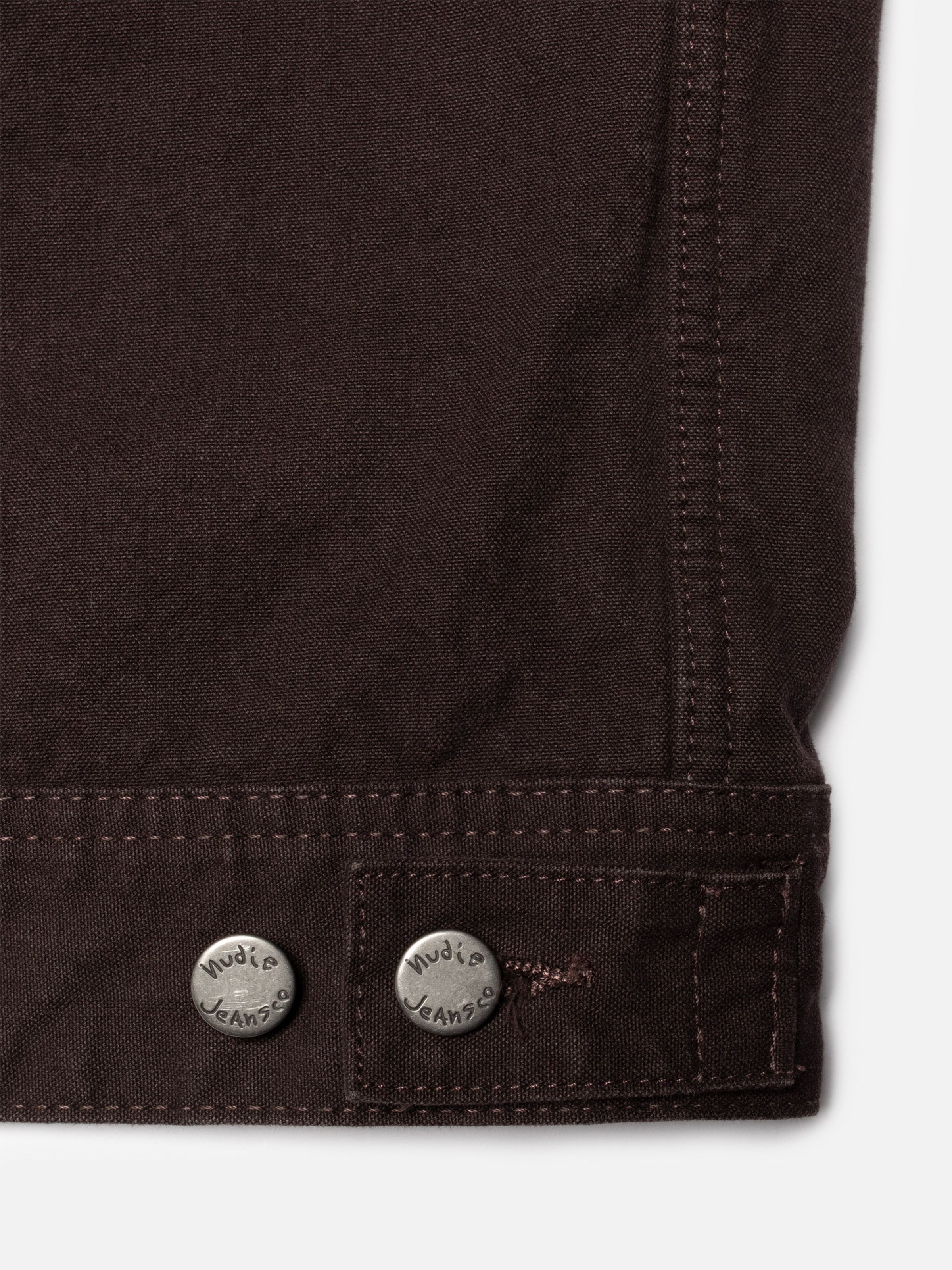 Nudie Jeans Blake Zip Jacket Dark Brown aus Bio-Baumwoll-Duck-Canvas, Detailaufnahme der silbernen Logo-Knöpfe am Saum.