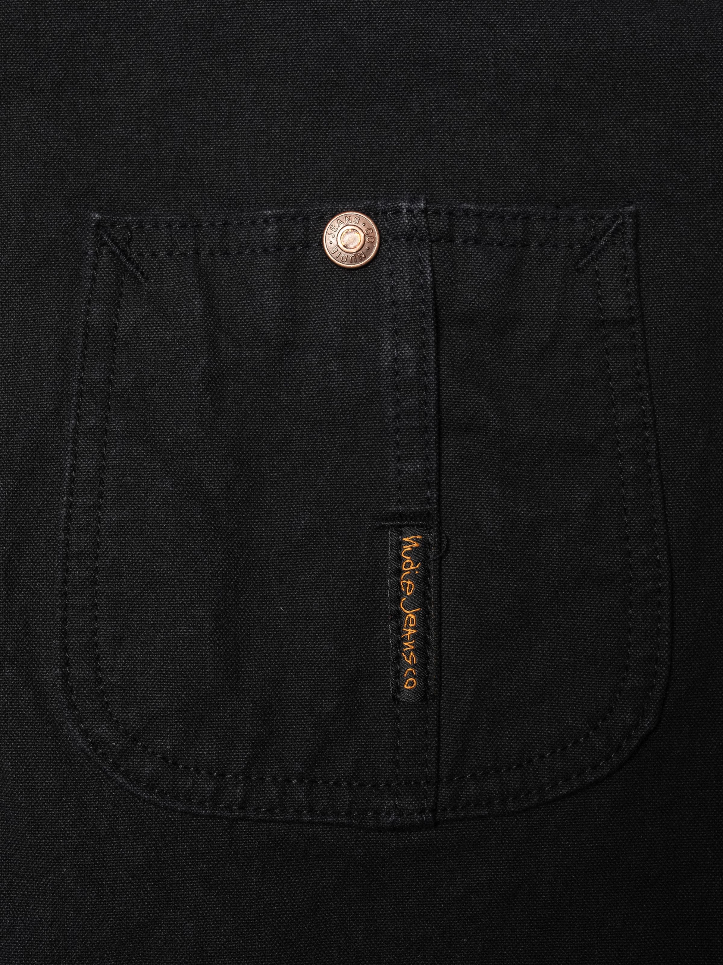 Nudie Jeans Howie Chore Jacket Duck Canvas in Schwarz aus Bio-Baumwolle, Nahaufnahme von Brusttasche und orangem Logo-Tab.