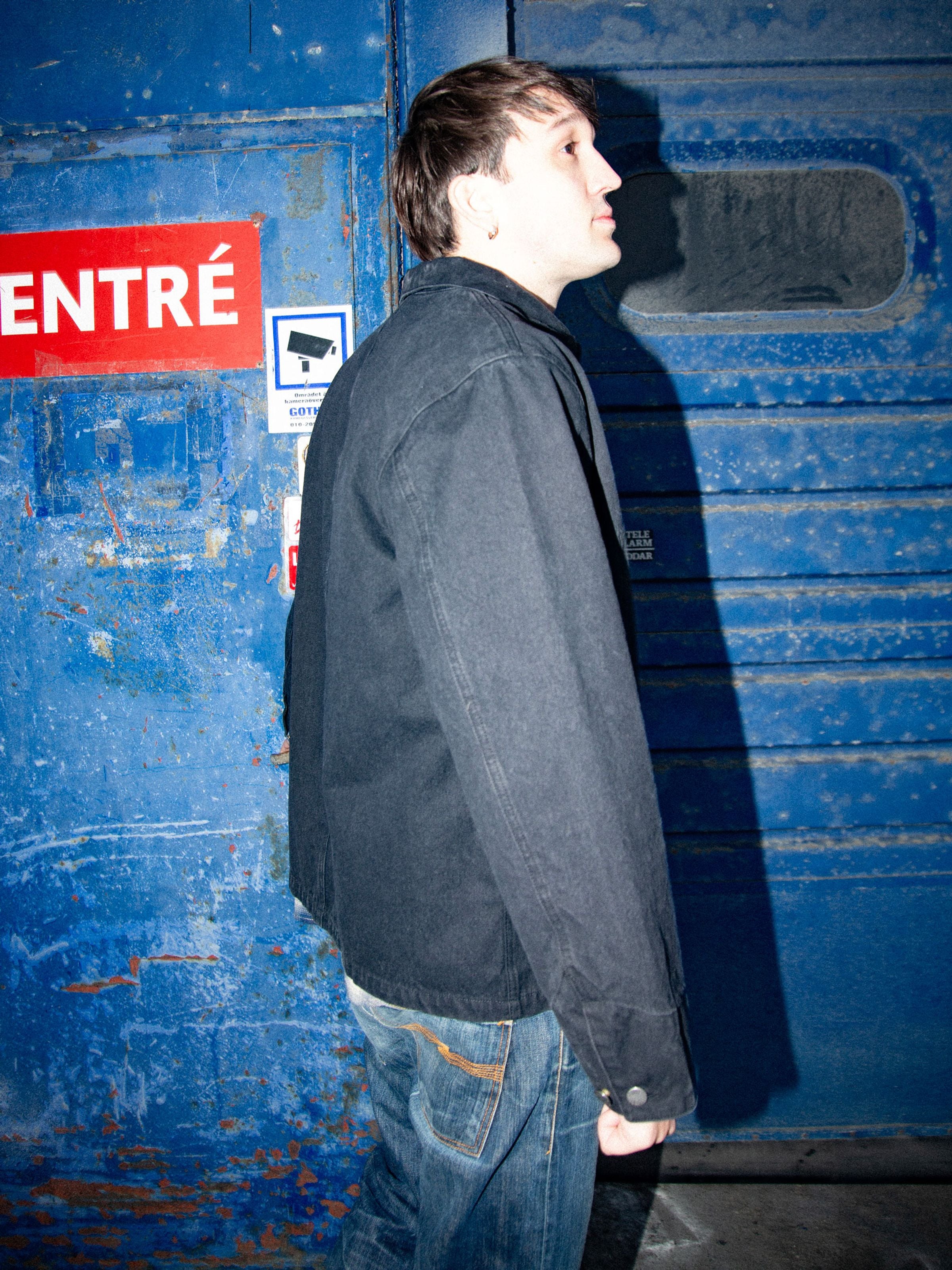 Nudie Jeans Howie Chore Jacket aus schwarzem Bio-Baumwoll-Duck-Canvas, Seitenansicht am Model vor blauer Industrietür.