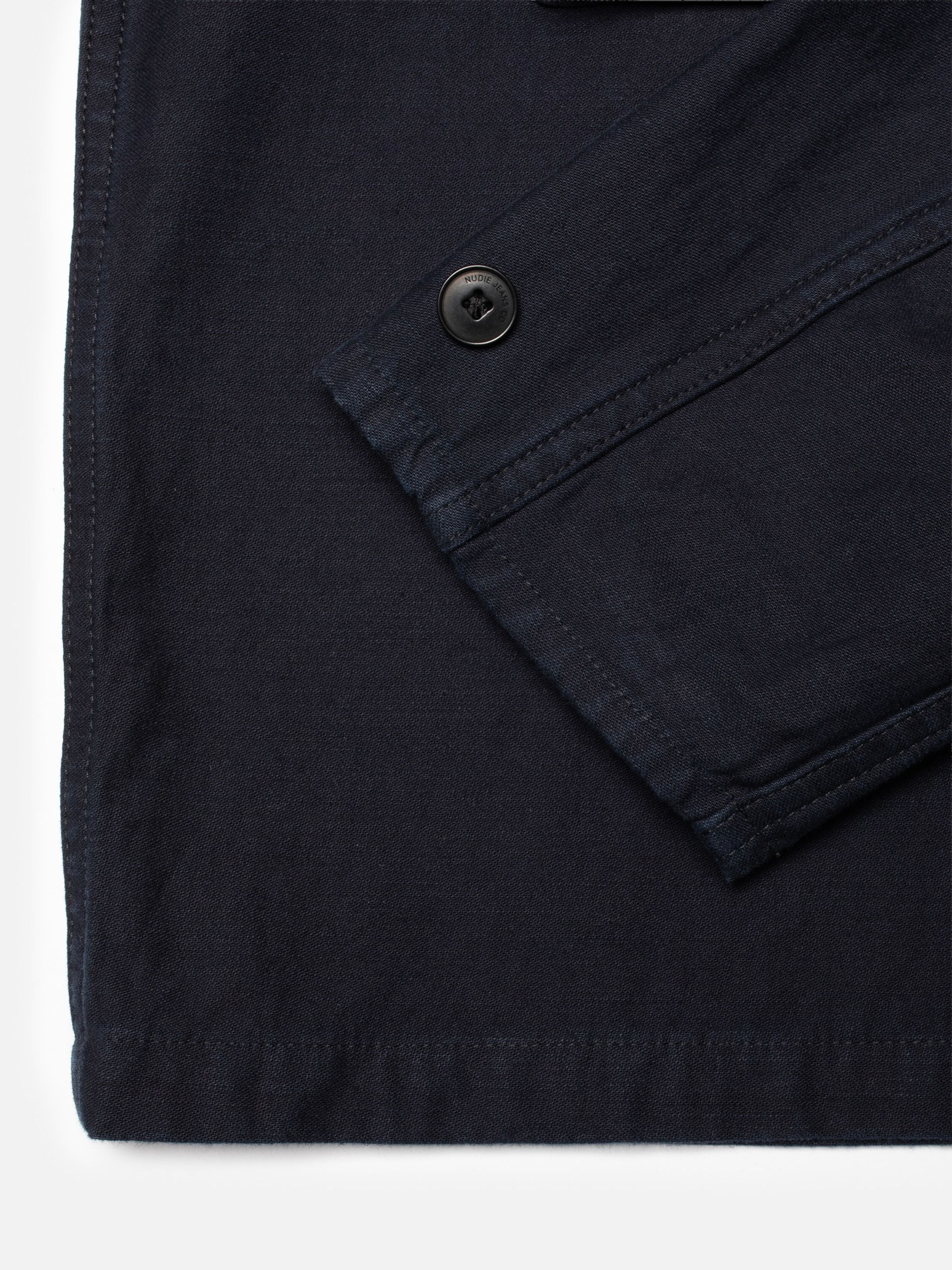 Nudie Jeans Nino Shirt Back Sateen Indigo, Nahaufnahme der Ärmelmanschette und des Markenknopfs auf Bio-Baumwolle.