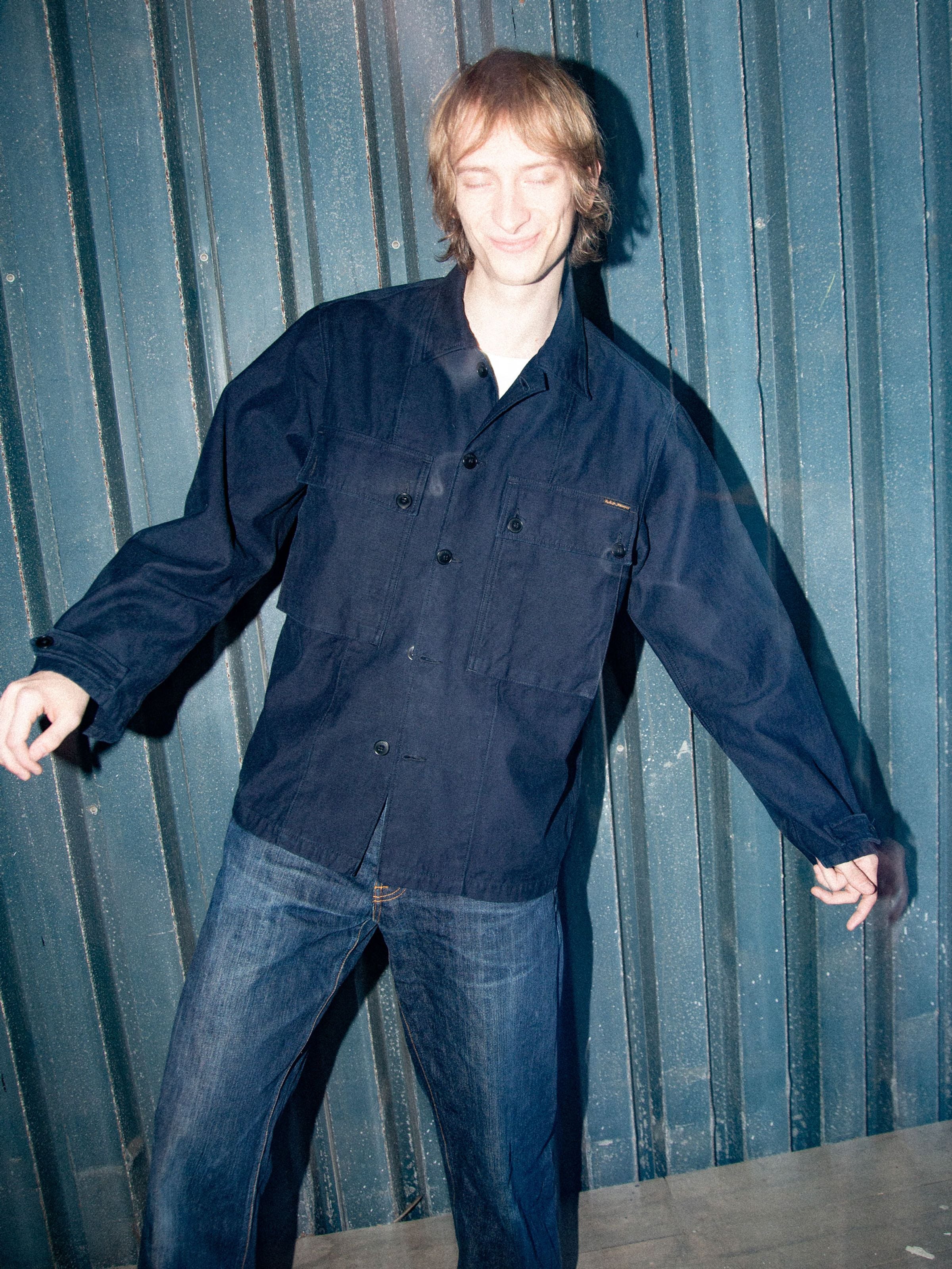 Nudie Jeans Nino Shirt Back Sateen Indigo, ein Overshirt aus Bio-Baumwolle an einem Model vor einer Metallwand.