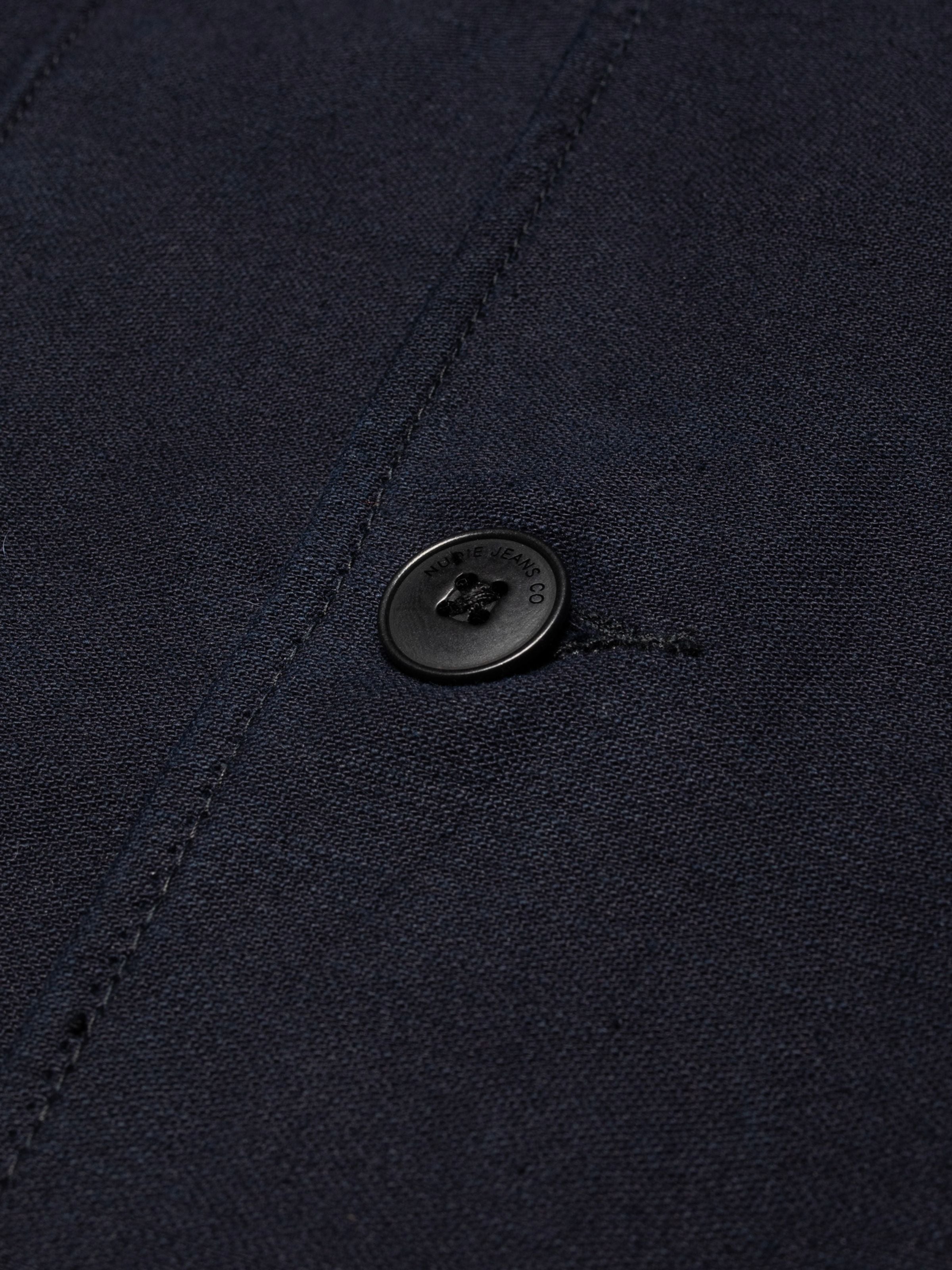 Nudie Jeans Nino Shirt Back Sateen Indigo, Nahaufnahme der Textur aus Bio-Baumwolle und des gravierten Knopfes.
