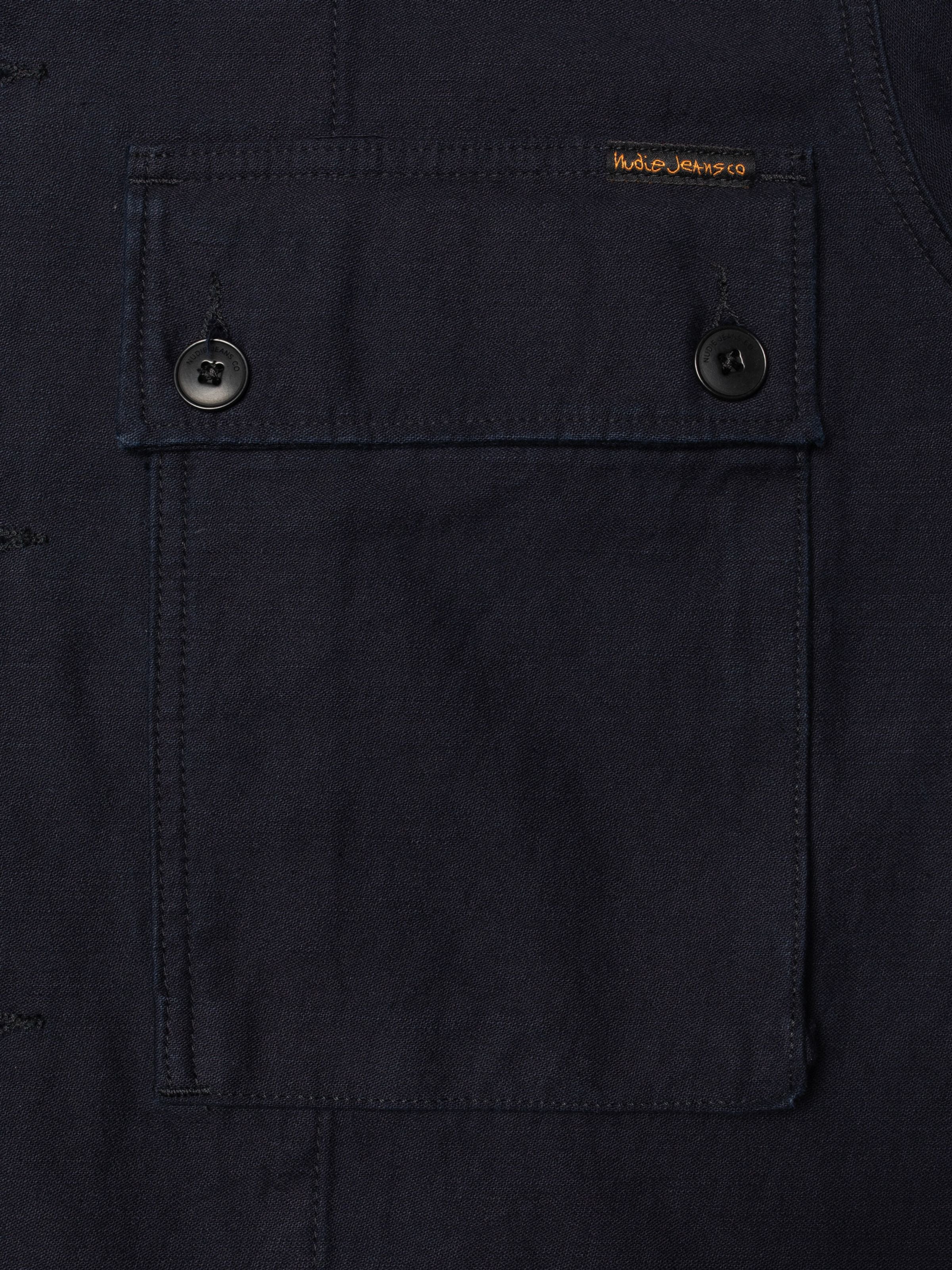 Nudie Jeans Nino Shirt Back Sateen Indigo, Nahaufnahme der geknöpften Brusttasche aus Bio-Baumwolle mit orangem Logo-Label.