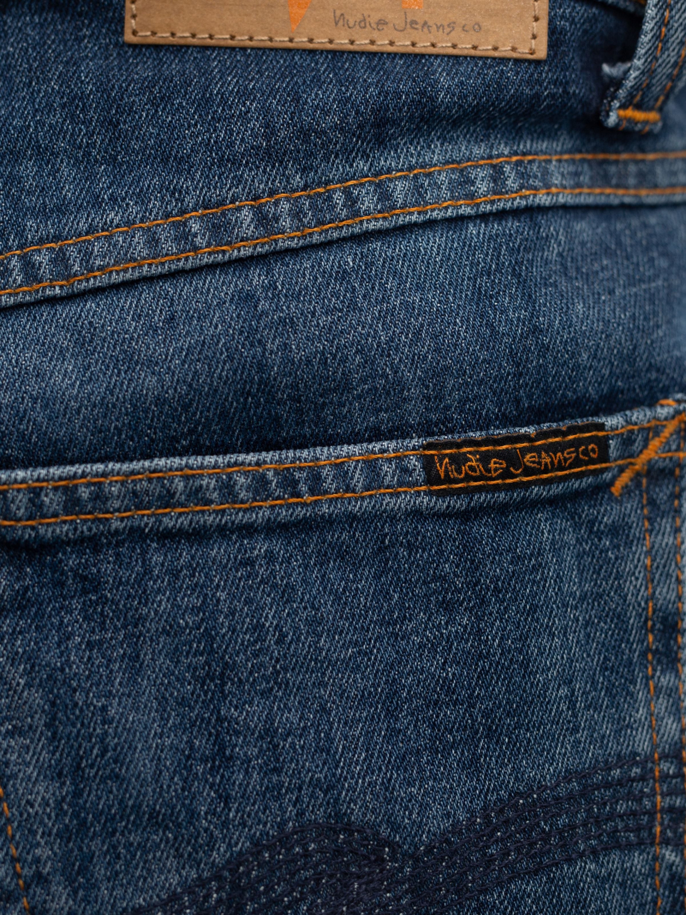 Nudie Jeans Solid Ollie Blue Echo Tracks, a close-up of orange stitching and black logo tab on blue organic cotton denim.