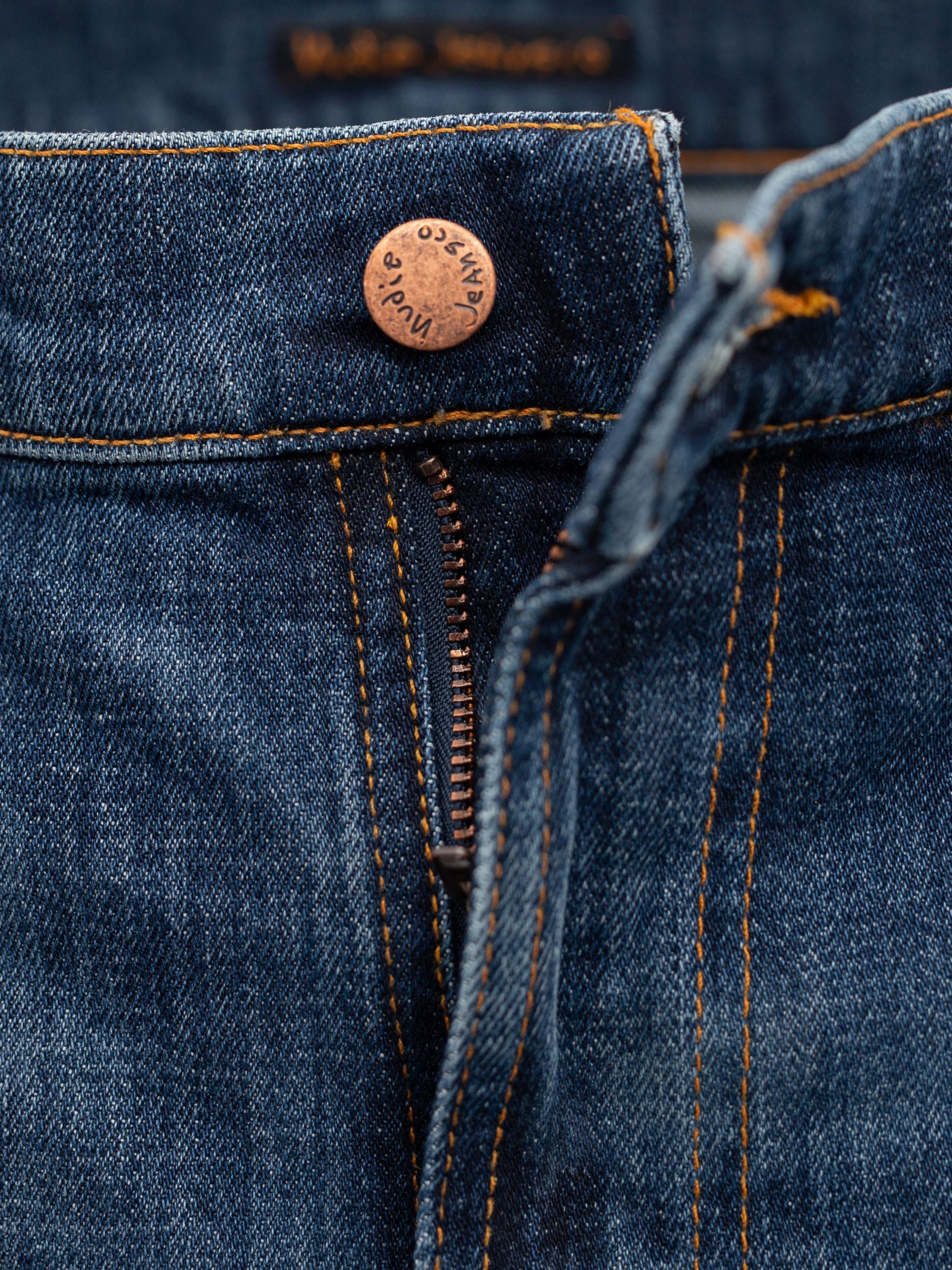 Nudie Jeans Lean Dean Echo Tracks, a close-up of the zip fly and copper button on dark blue washed organic cotton denim.