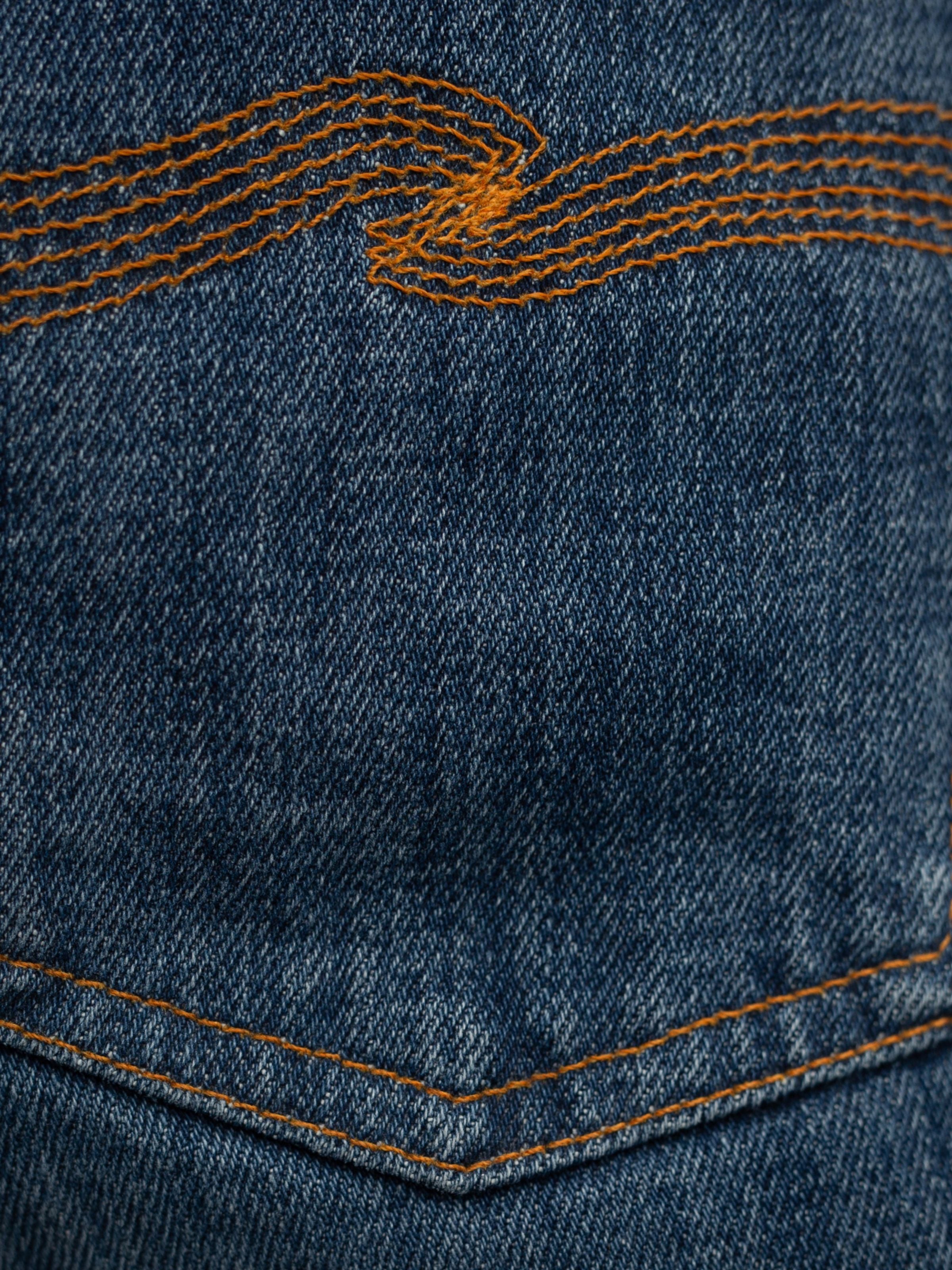 Nudie Jeans Steady Eddie II Twisted Tracks, Nahaufnahme von dunkelblauem Denim aus Bio-Baumwolle und oranger Stickerei.