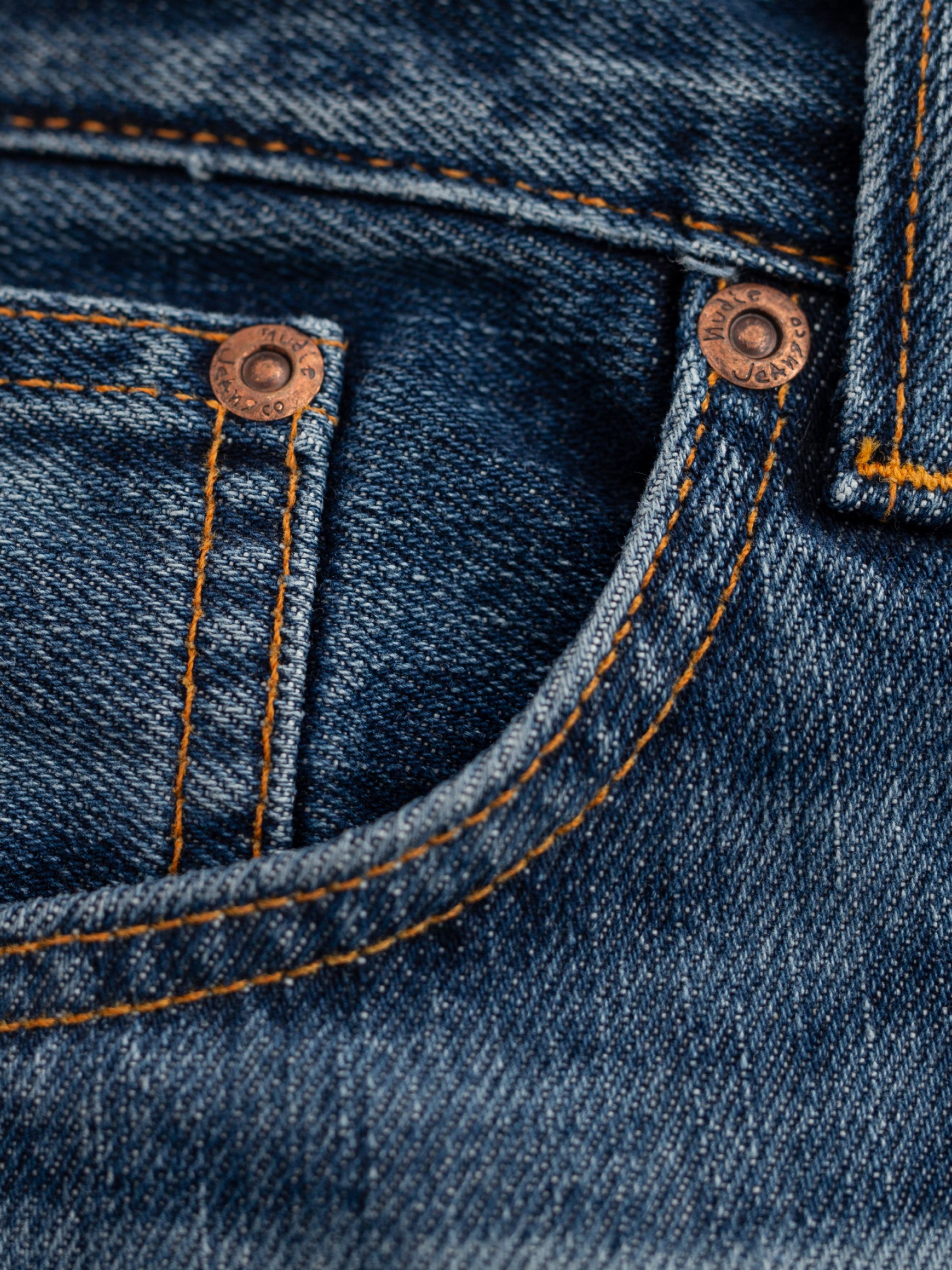 Nudie Jeans Steady Eddie II Twisted Tracks, Nahaufnahme von Bio-Baumwoll-Denim mit orangefarbenen Nähten und Kupfernieten.