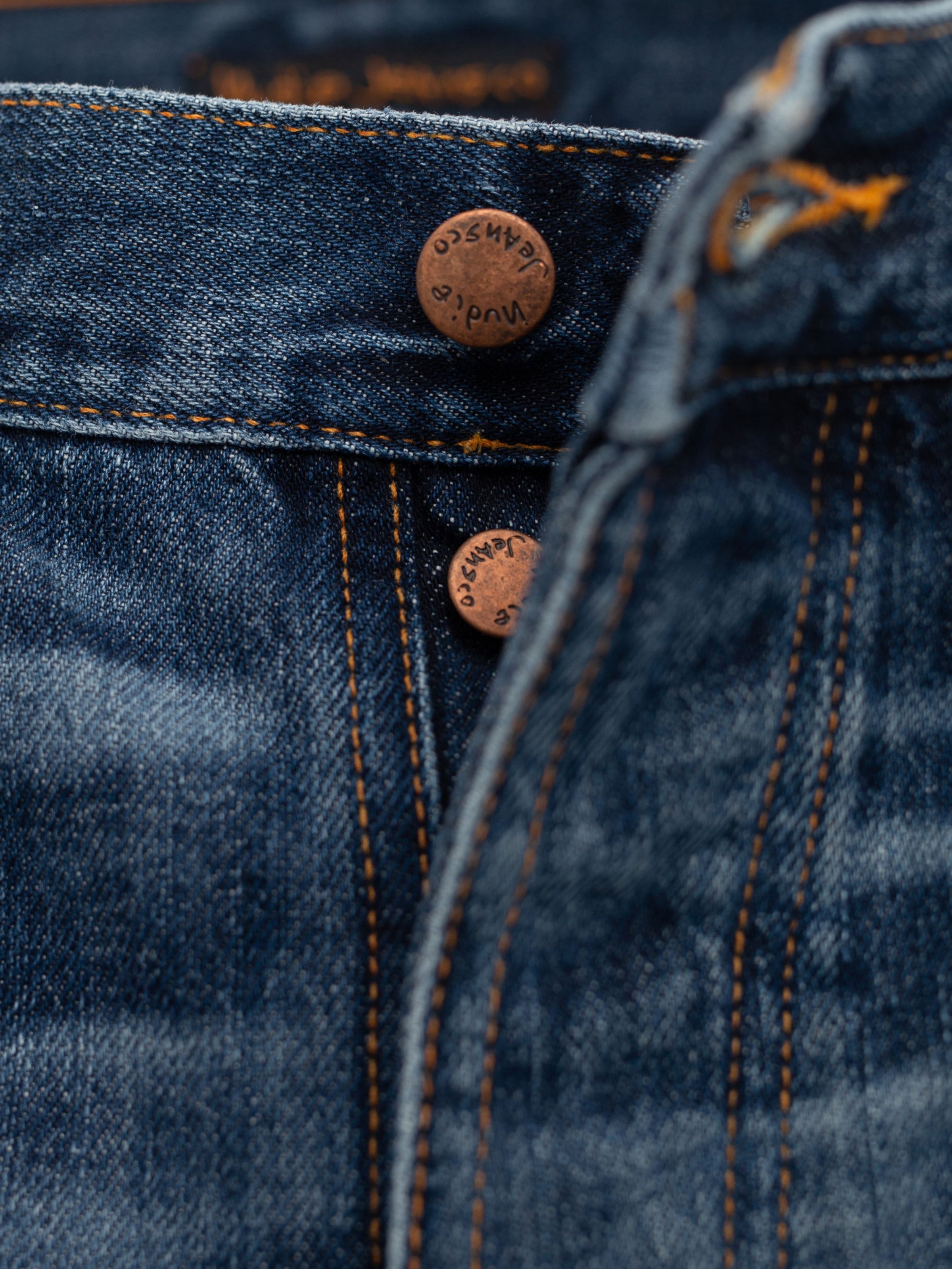 Nudie Jeans Steady Eddie II Twisted Tracks, Nahaufnahme der Kupferknöpfe und orangefarbenen Nähte auf Bio-Baumwoll-Denim.
