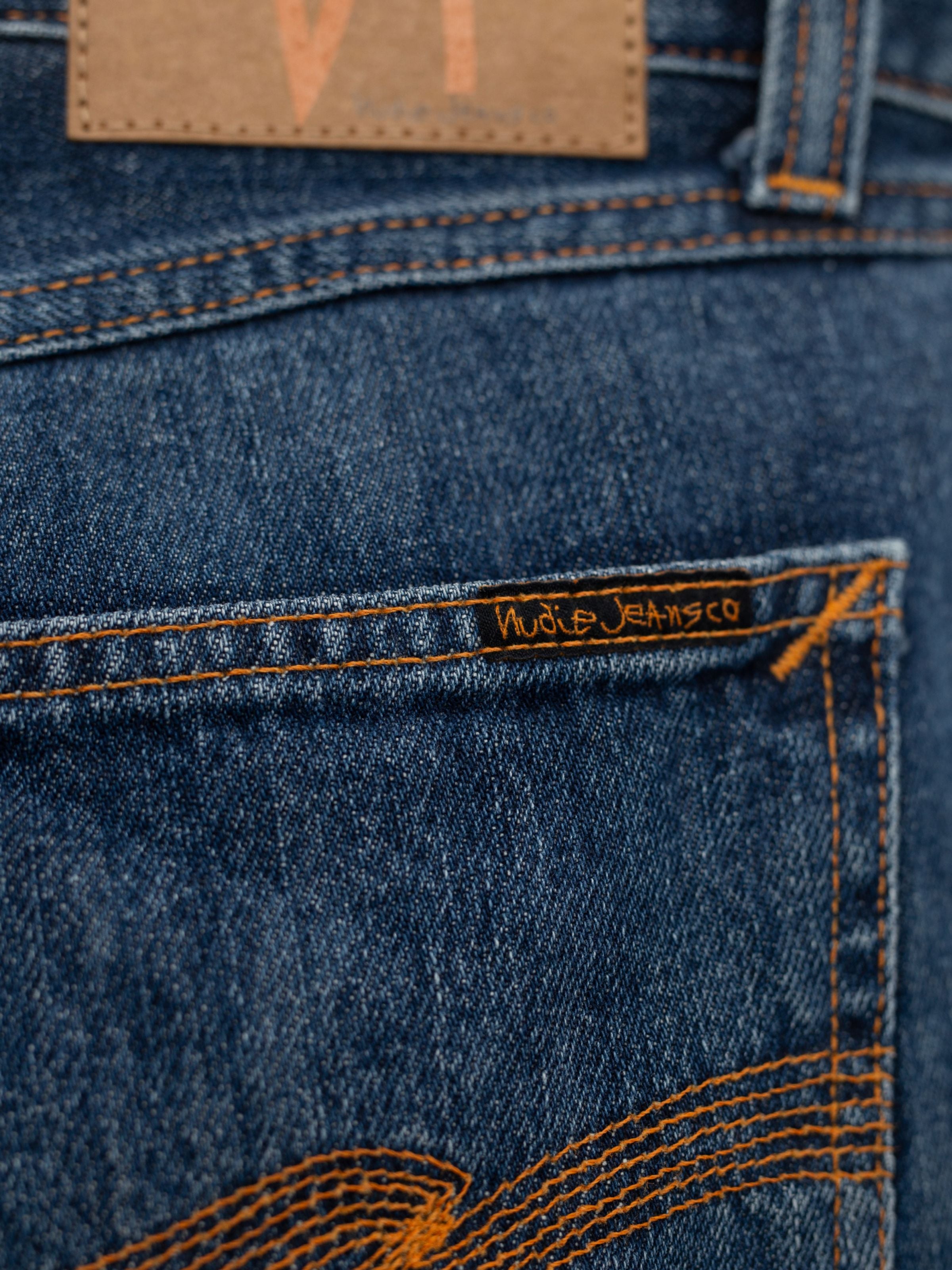 Nudie Jeans Steady Eddie II Twisted Tracks, Nahaufnahme des Gesäßtaschen-Labels auf blauem Rigid Denim aus Bio-Baumwolle.