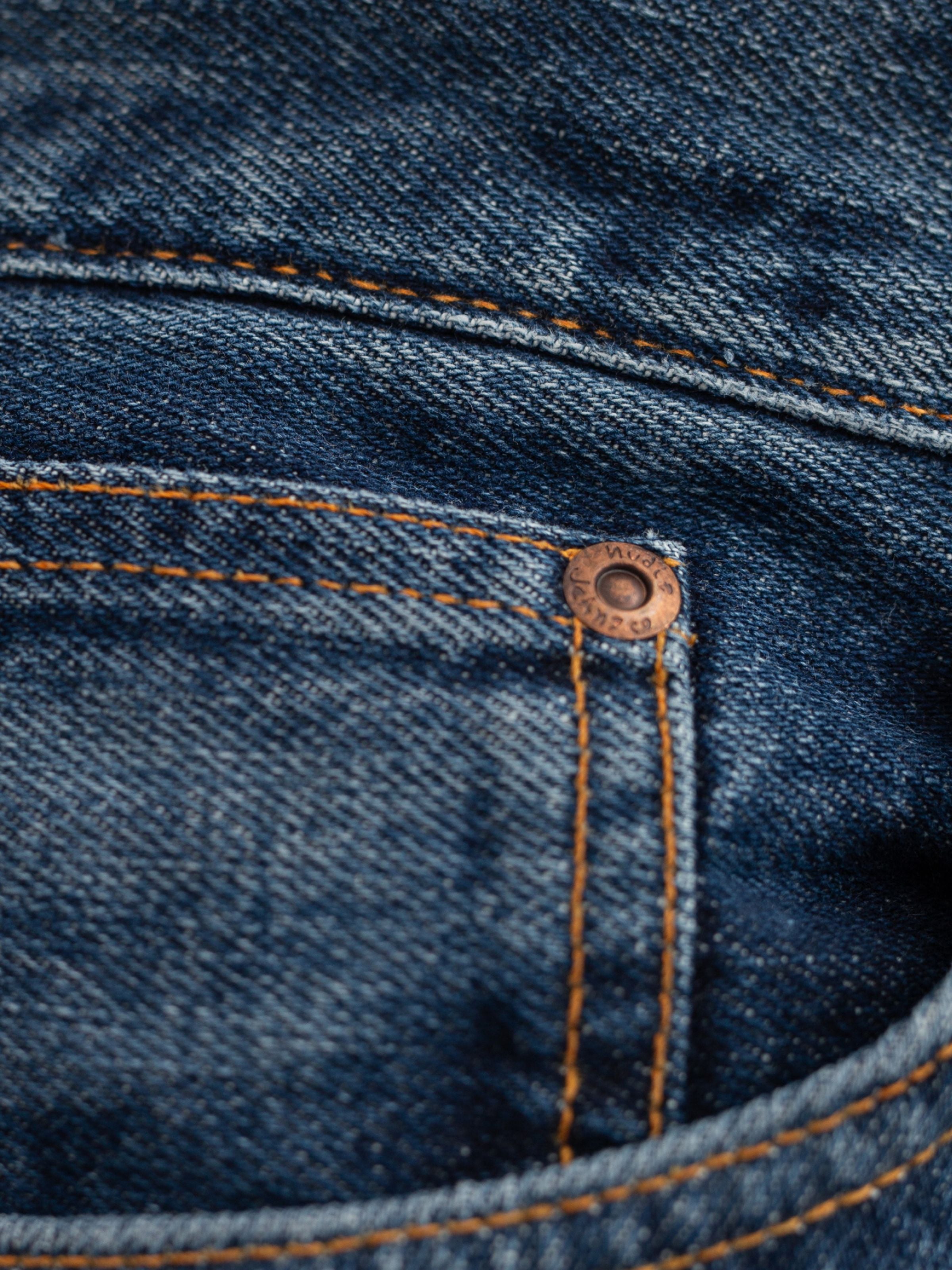 Nudie Jeans Rad Rufus Blue Twisted Tracks, a close-up of organic cotton rigid denim with orange stitching and copper rivet.