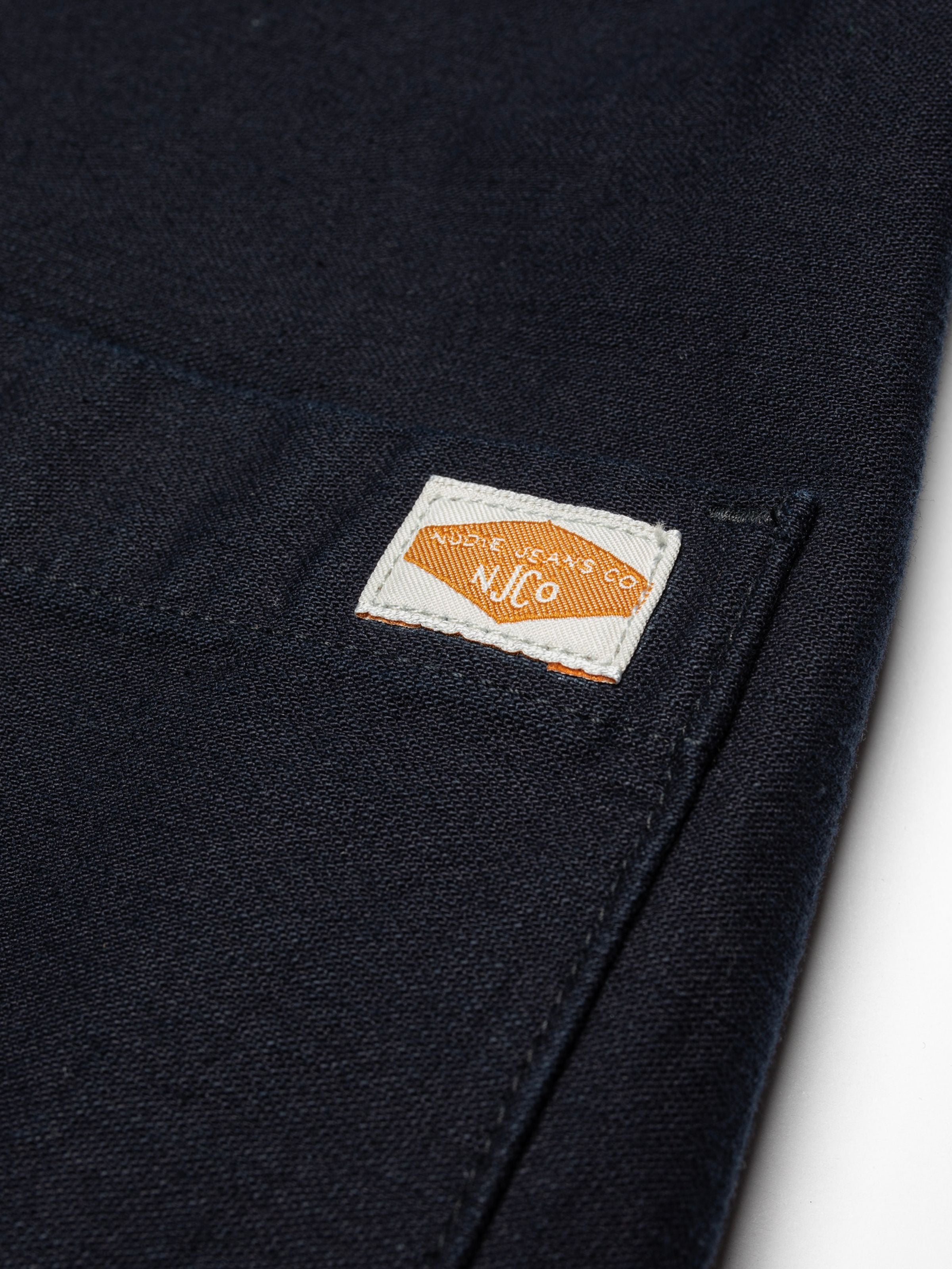Nudie Jeans Lovis Jacket aus Bio-Baumwolle Back Sateen in Indigo, Nahaufnahme des gewebten NJCO Logo-Labels an der Tasche.