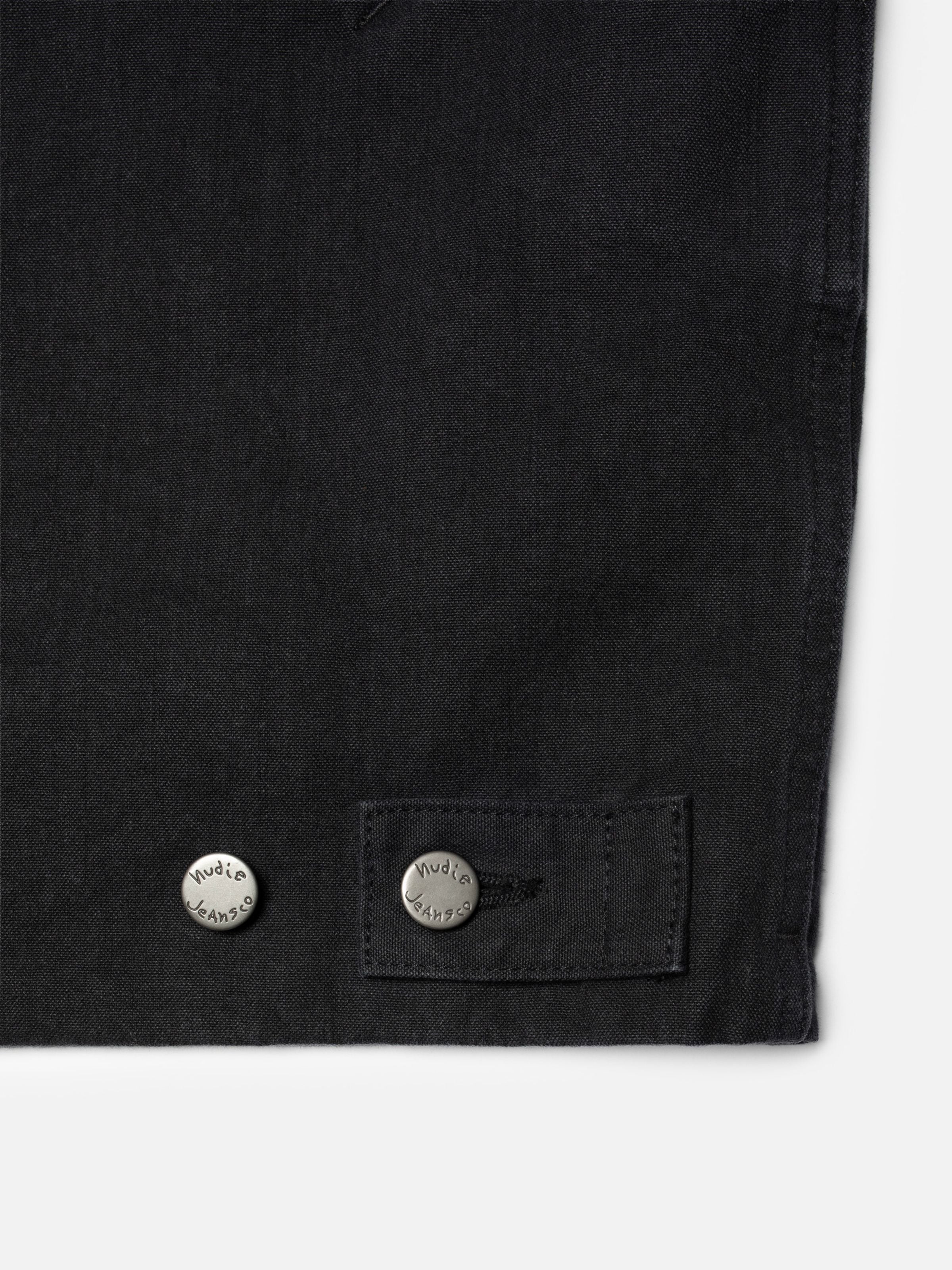 Nudie Jeans Lana Jacket in black organic cotton duck canvas, close-up of the hem with engraved metal buttons.