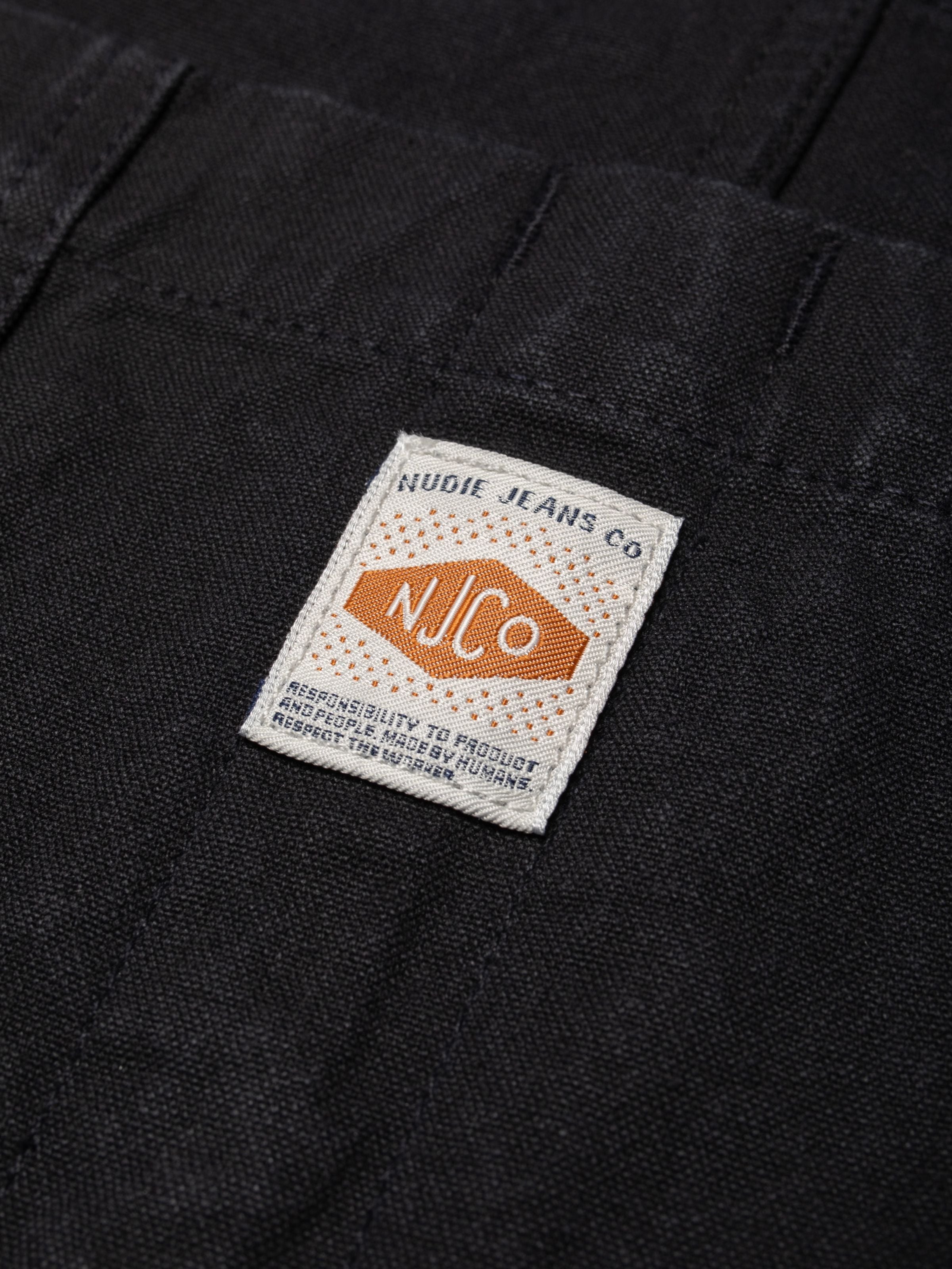 Nudie Jeans Klara Dungarees Duck Canvas, a close-up of the brand patch on the black organic cotton canvas fabric.