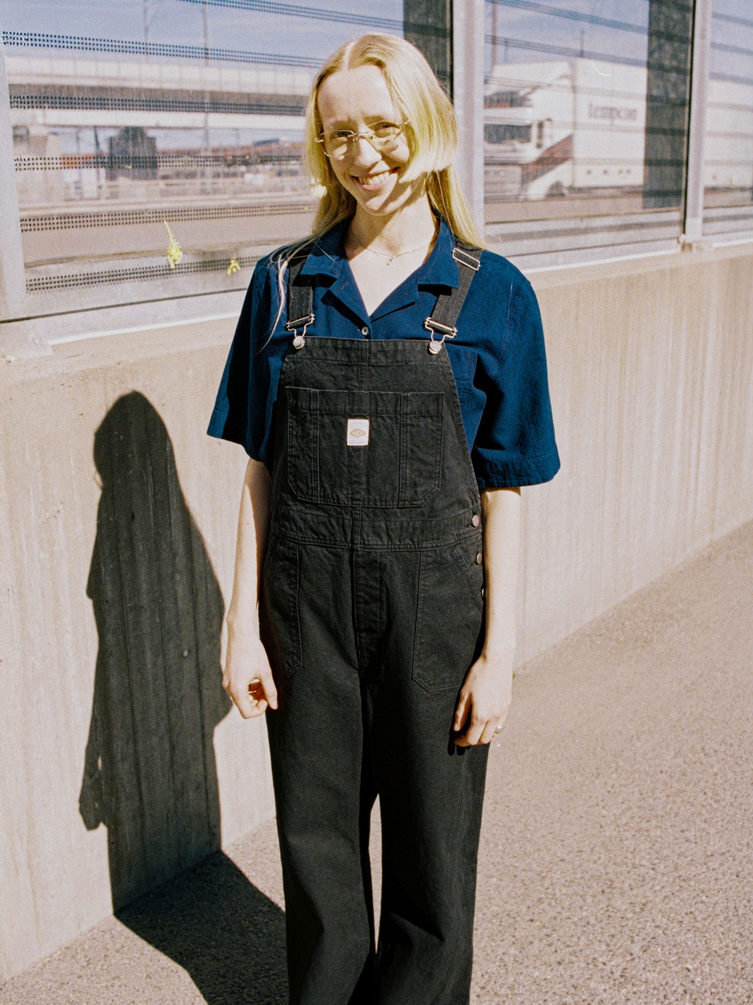 Nudie Jeans Klara Dungarees Duck Canvas in black, worn by a model outdoors in a regular fit style.