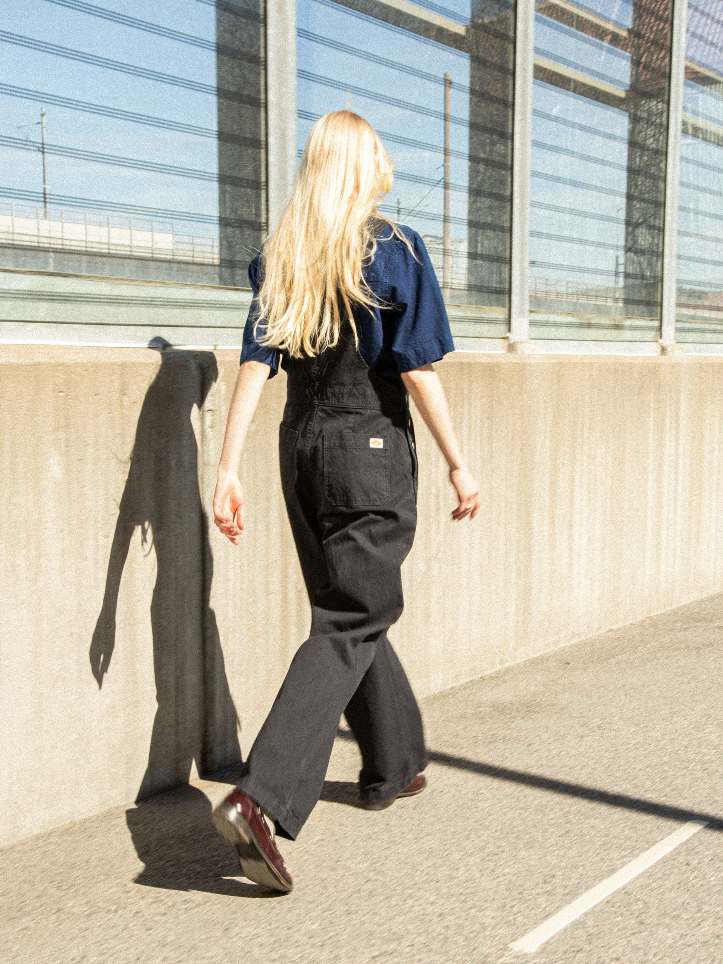 Nudie Jeans Klara Dungarees Duck Canvas in black, worn by a model walking outdoors and shown from the back.