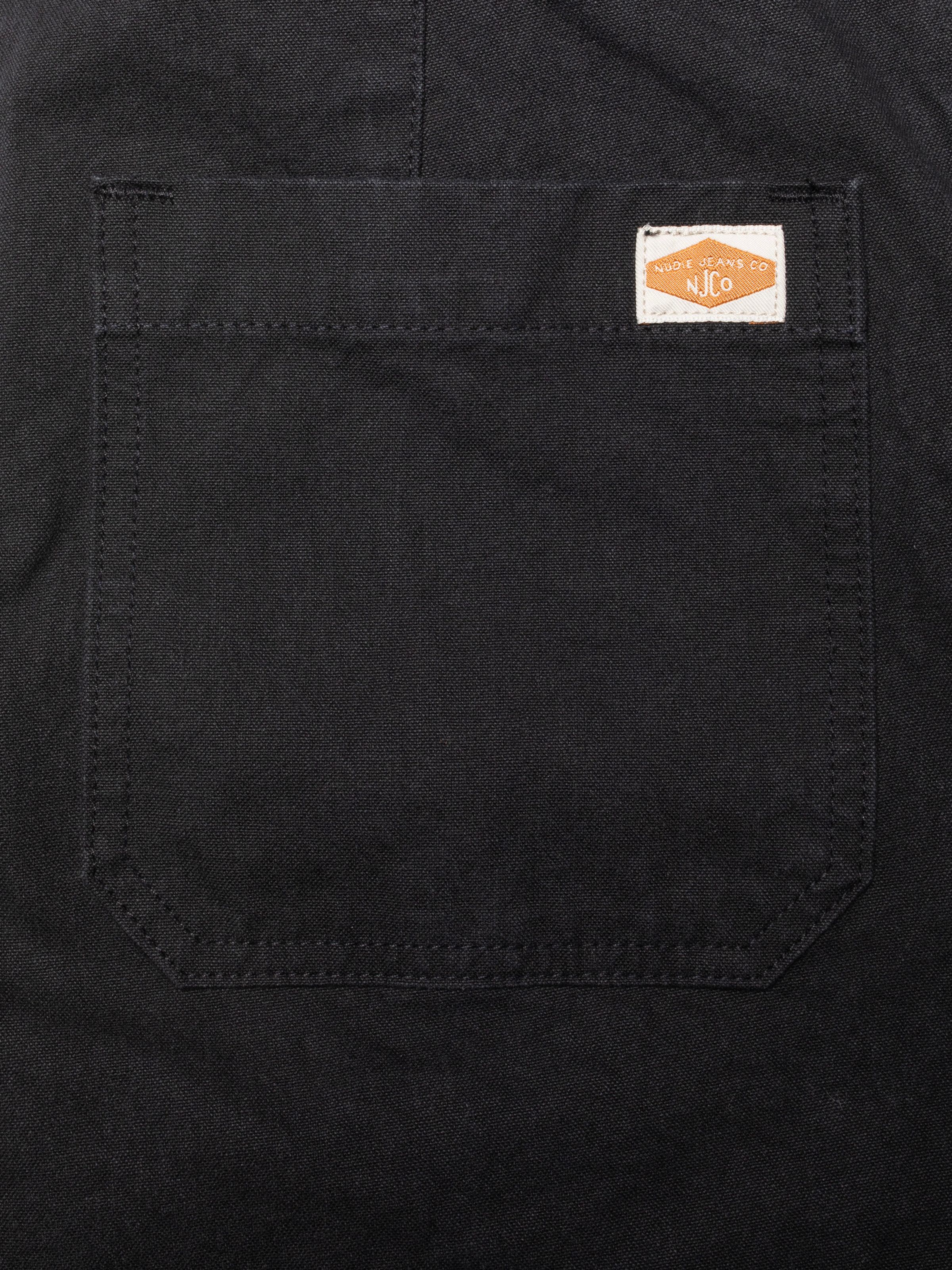 Nudie Jeans Klara Dungarees Duck Canvas, a close-up of the pitch-black duck canvas pocket with brand label.
