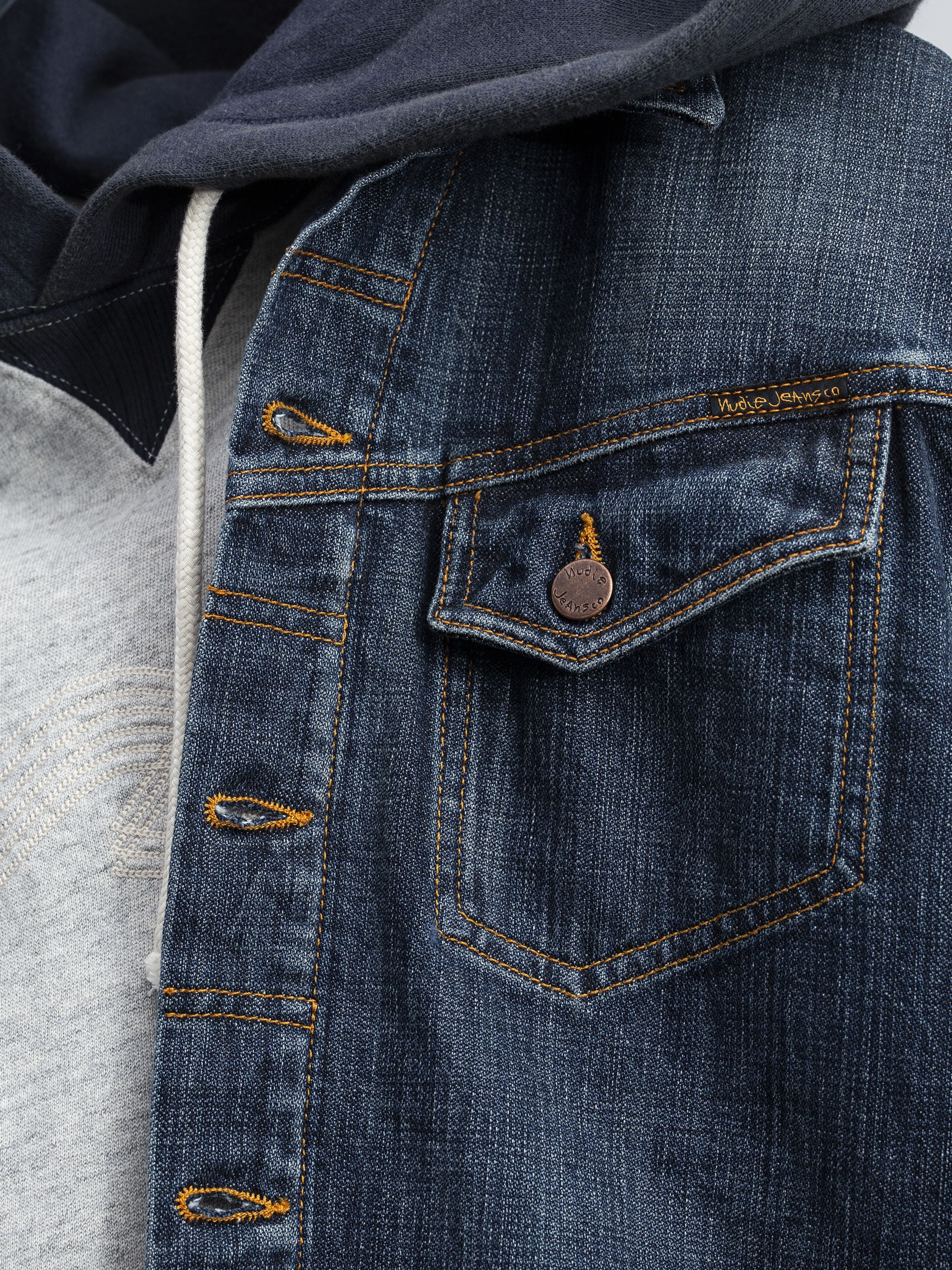 Nudie Jeans Conny Denim Jacket Big Wave, Nahaufnahme des Denims aus Bio-Baumwolle mit orangefarbenen Nähten und Kupferknopf.