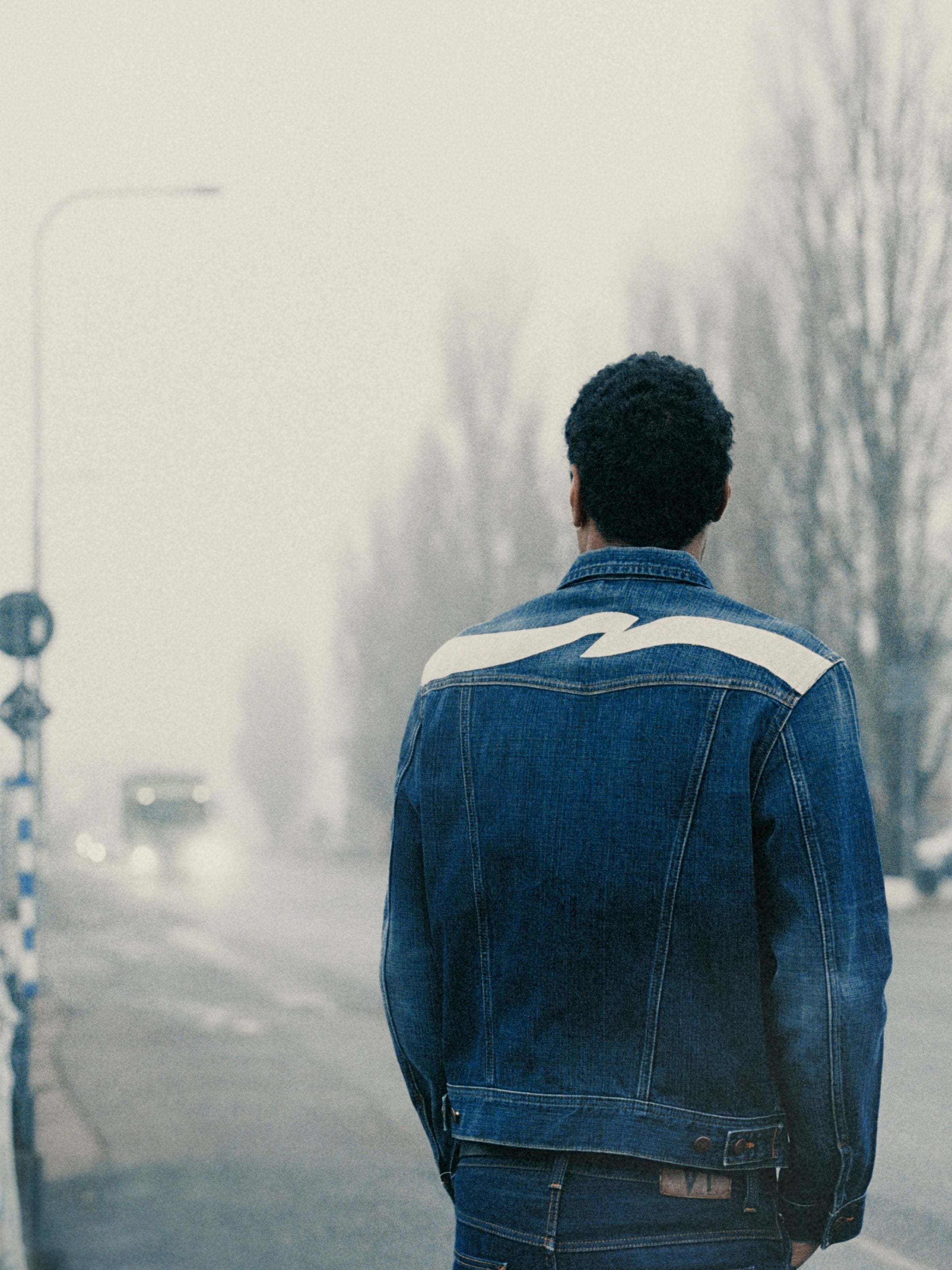 Nudie Jeans Conny Jeansjacke Big Wave aus mittelblauer Bio-Baumwolle mit weißer Rückenstickerei auf einer nebligen Straße.