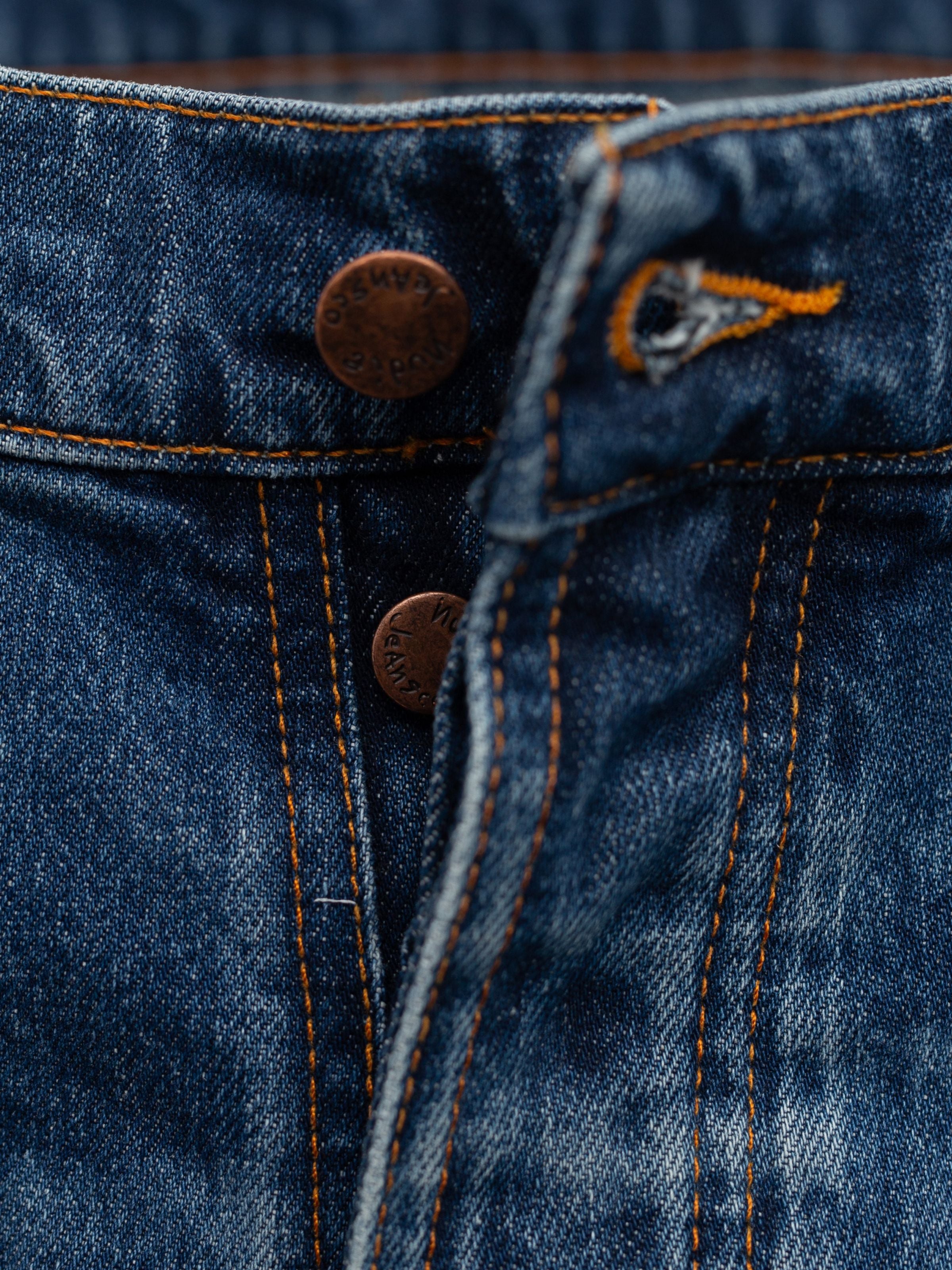 Nudie Jeans Grim Tim Life Tracks, a close-up of the copper button fly and orange stitching on mid blue organic cotton denim.