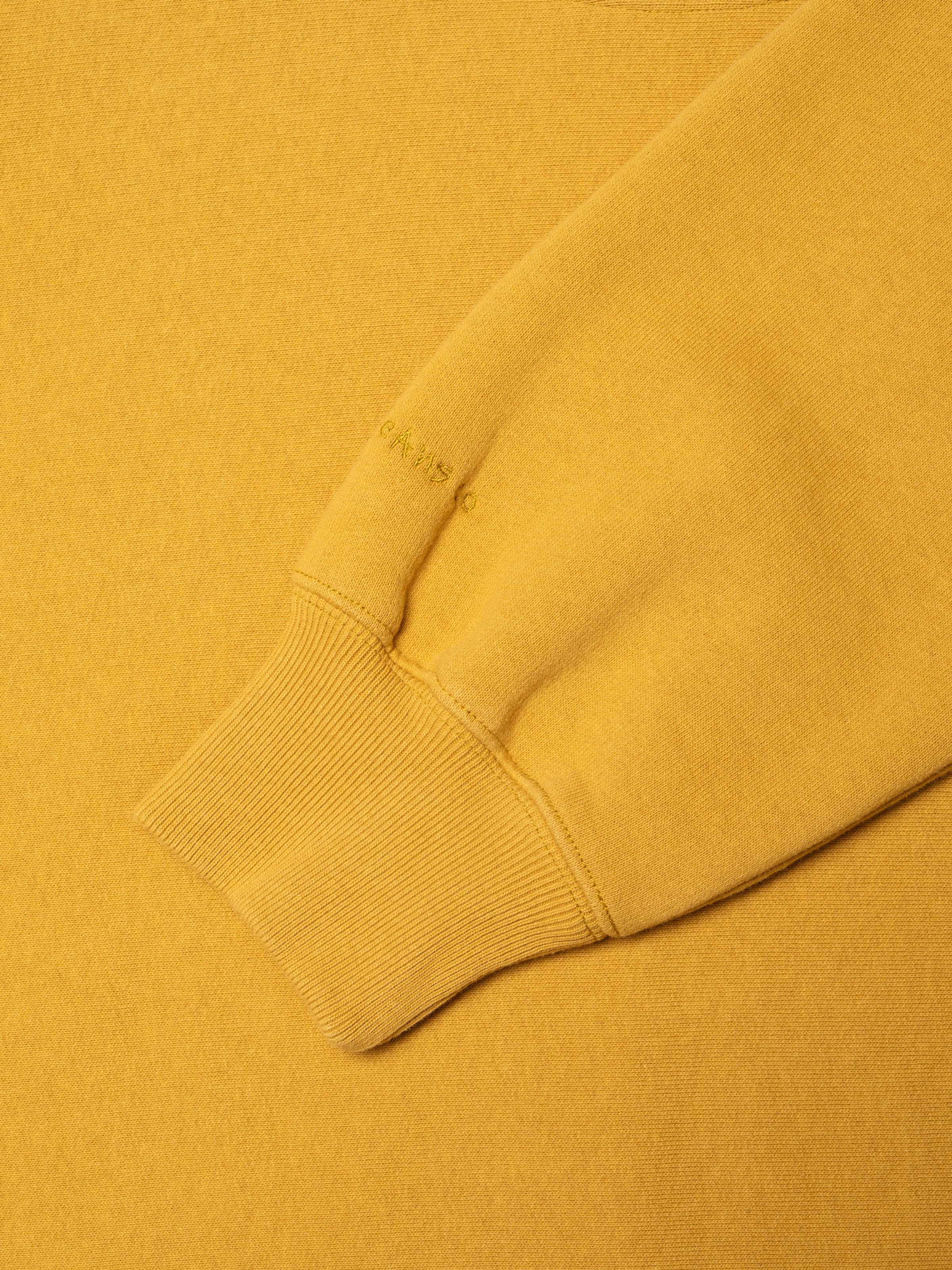 Nudie Jeans Hasse Crew Neck Sweatshirt in Old Yellow, showing a close-up of the sleeve cuff and logo embroidery.