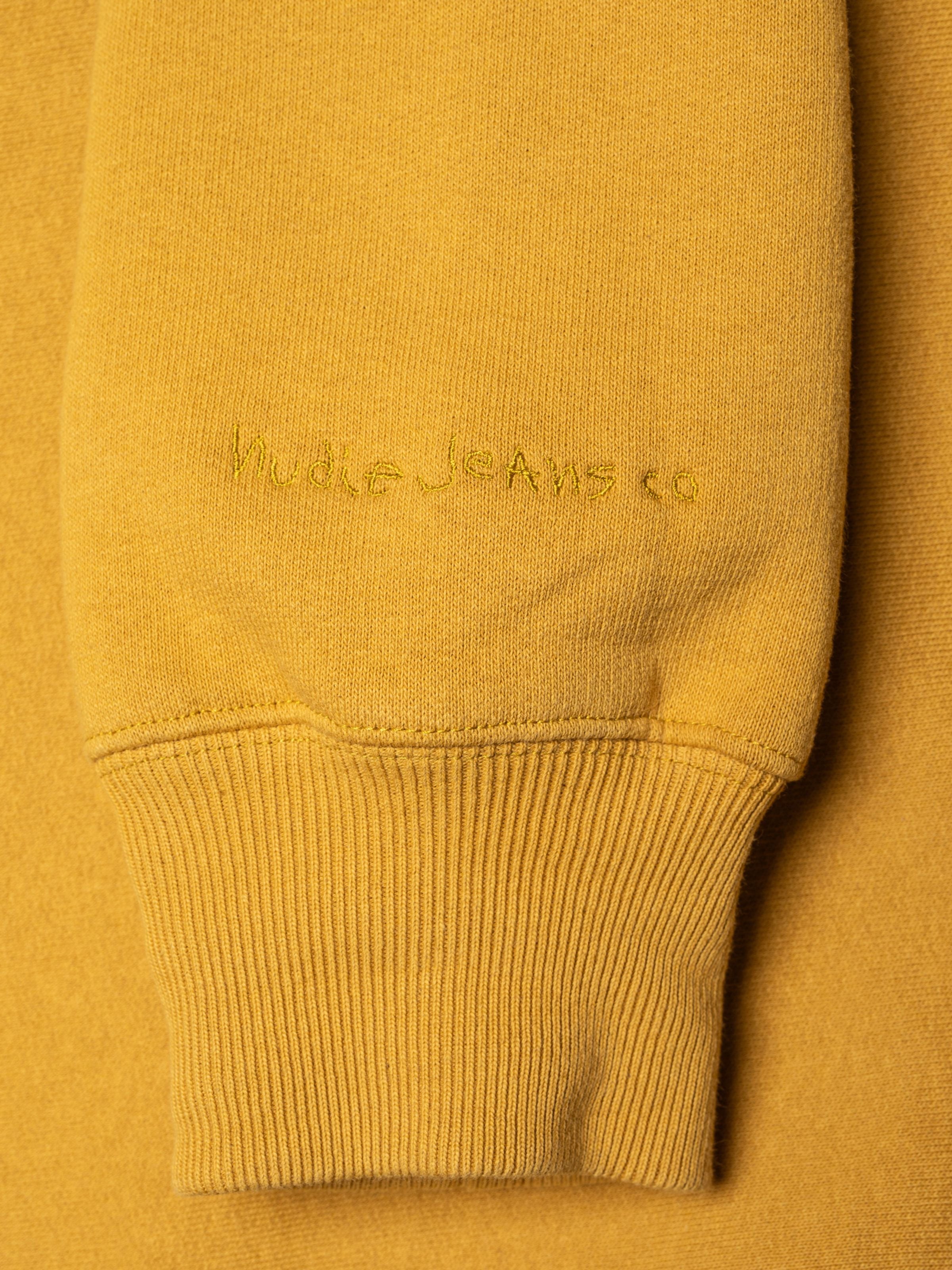 Nudie Jeans Hasse Crew Neck Sweatshirt in old yellow, close-up of the logo embroidery on the sleeve and heavy jersey texture.