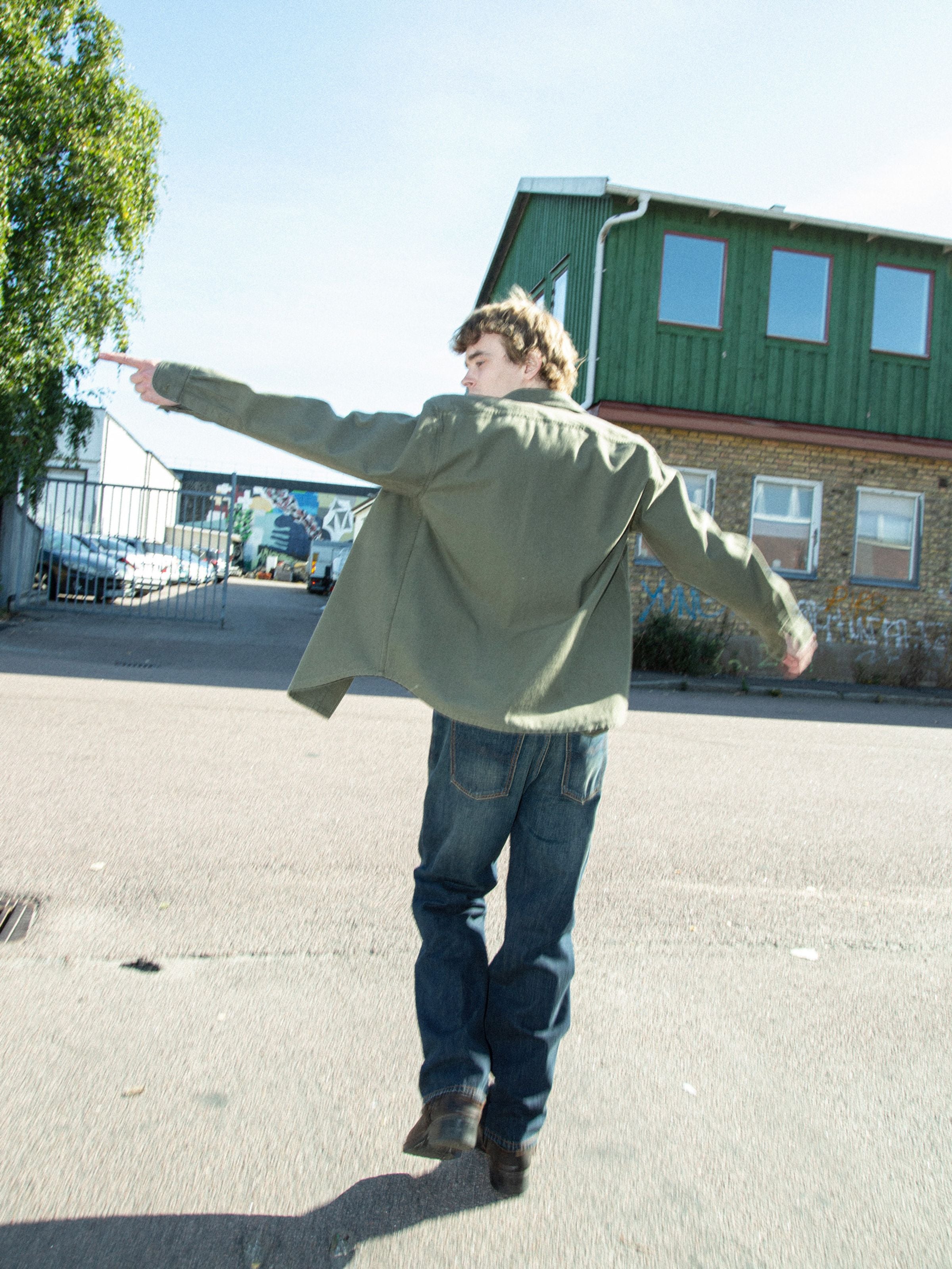 Nudie Jeans Julian Shirt in Olive Green, worn by a man from the back outside against an industrial backdrop.