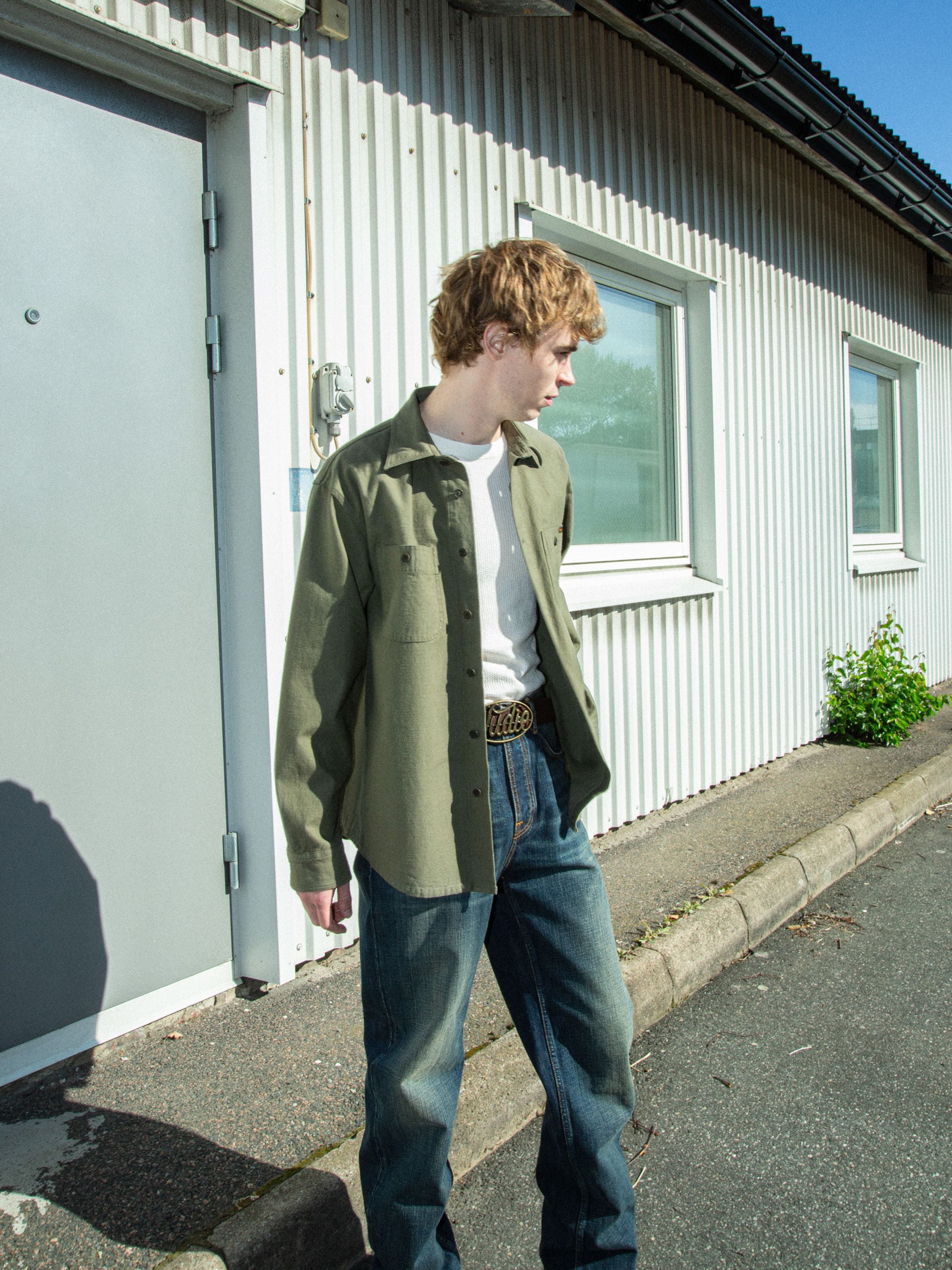 Nudie Jeans Julian Shirt Back Sateen in Olive Green, worn outdoors with blue jeans over a white t-shirt.