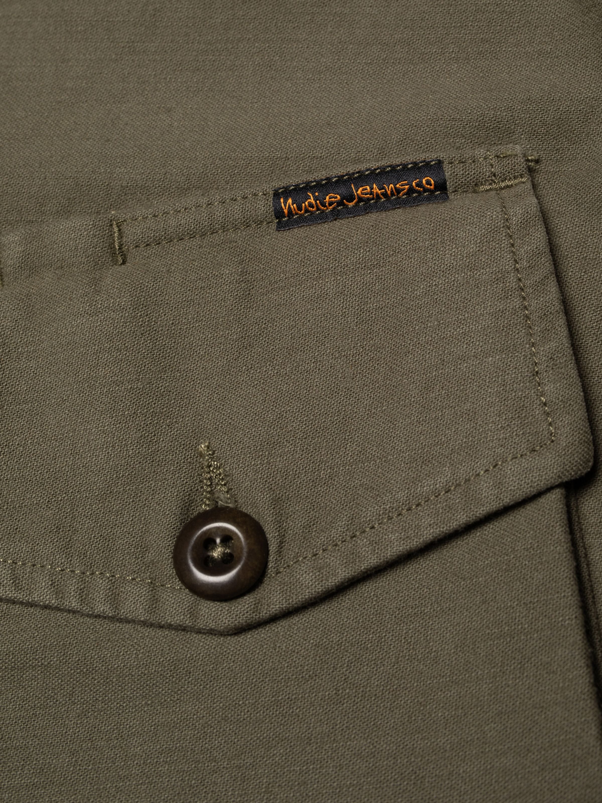 Nudie Jeans Bror Field Jacket in Olive Green, close-up of the chest pocket, corozo button, and organic cotton sateen texture.