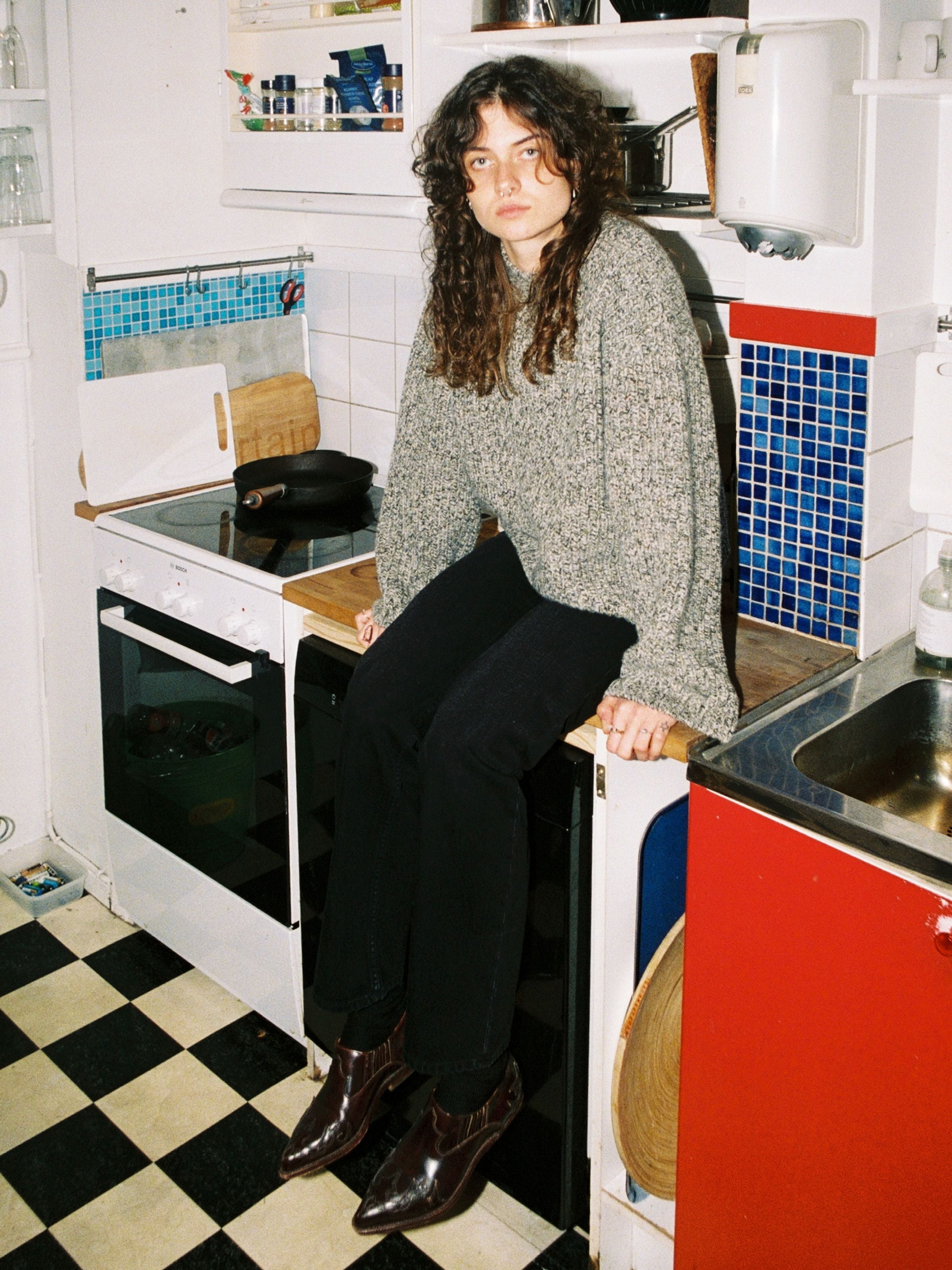 Nudie Jeans Gittan Sweater Chunky Knit in Green Melange, lifestyle shot of a model sitting on a kitchen counter.
