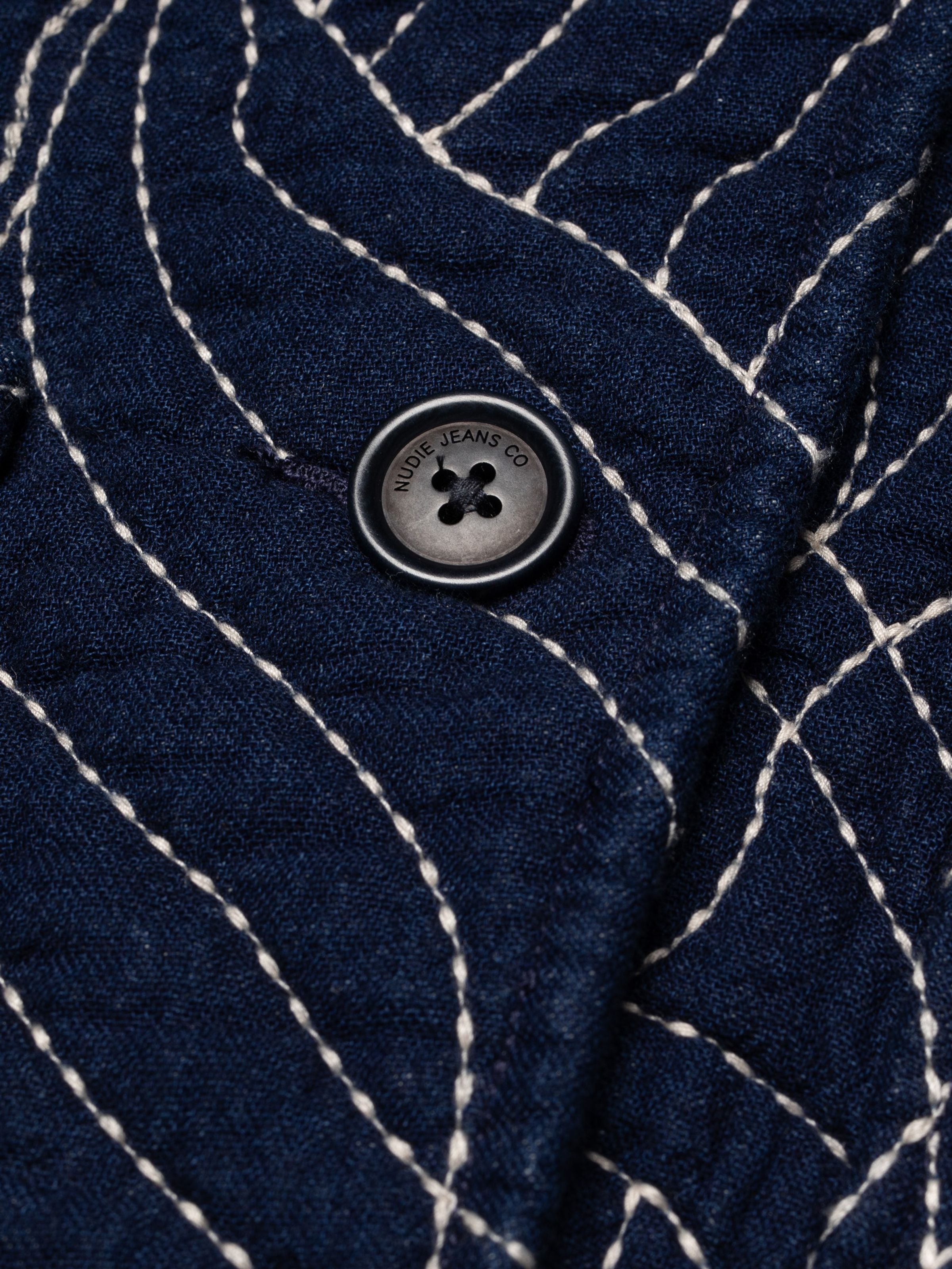 Nudie Jeans Lena Jacket Indigo Quilt, Nahaufnahme des gesteppten Indigostoffs mit geschwungenen Nähten und Corozoknopf.