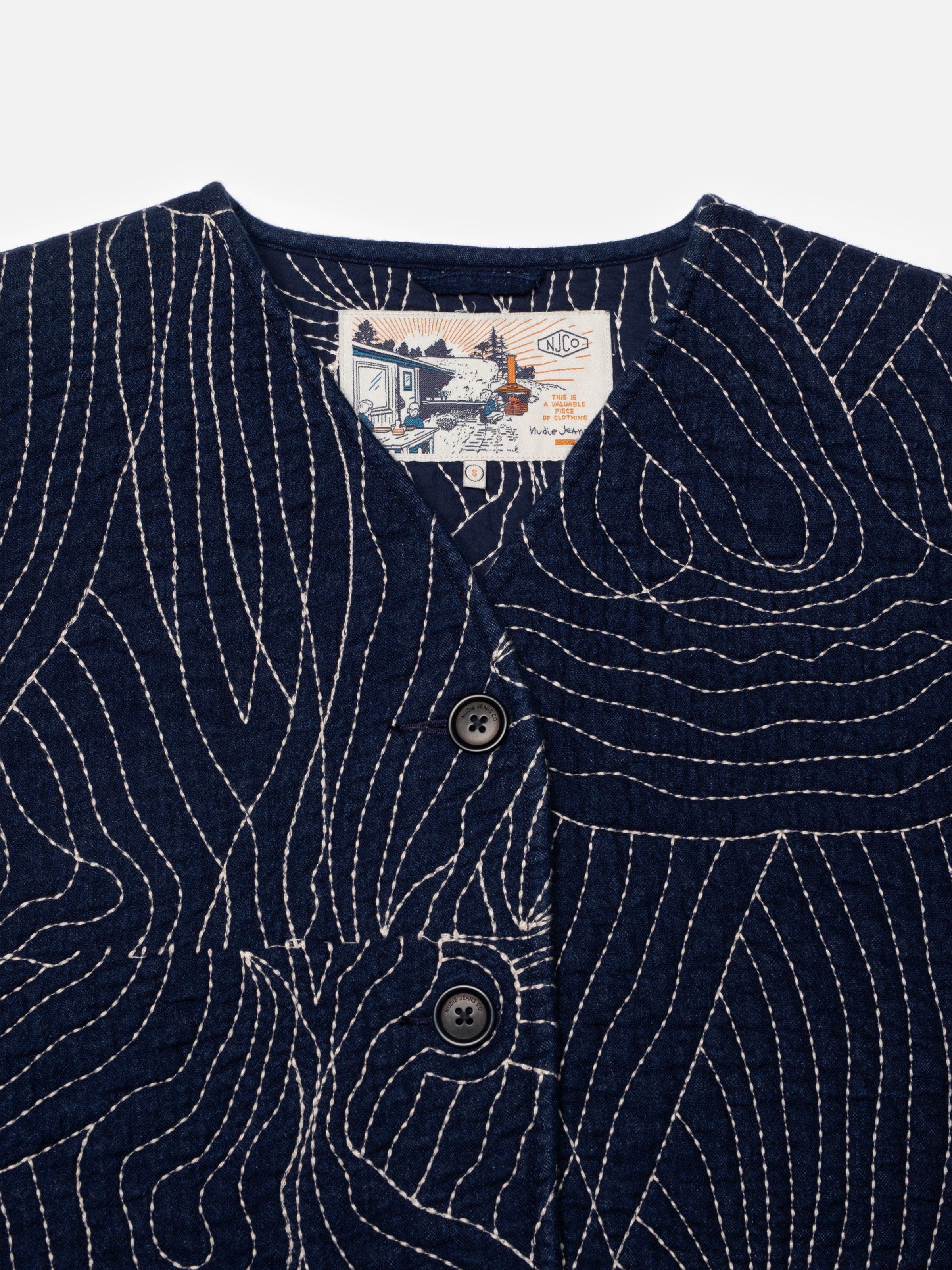 Nudie Jeans Lena Jacket Indigo Quilt, Nahaufnahme von blauem Steppstoff mit geschwungenen Steppmustern und Knöpfen.