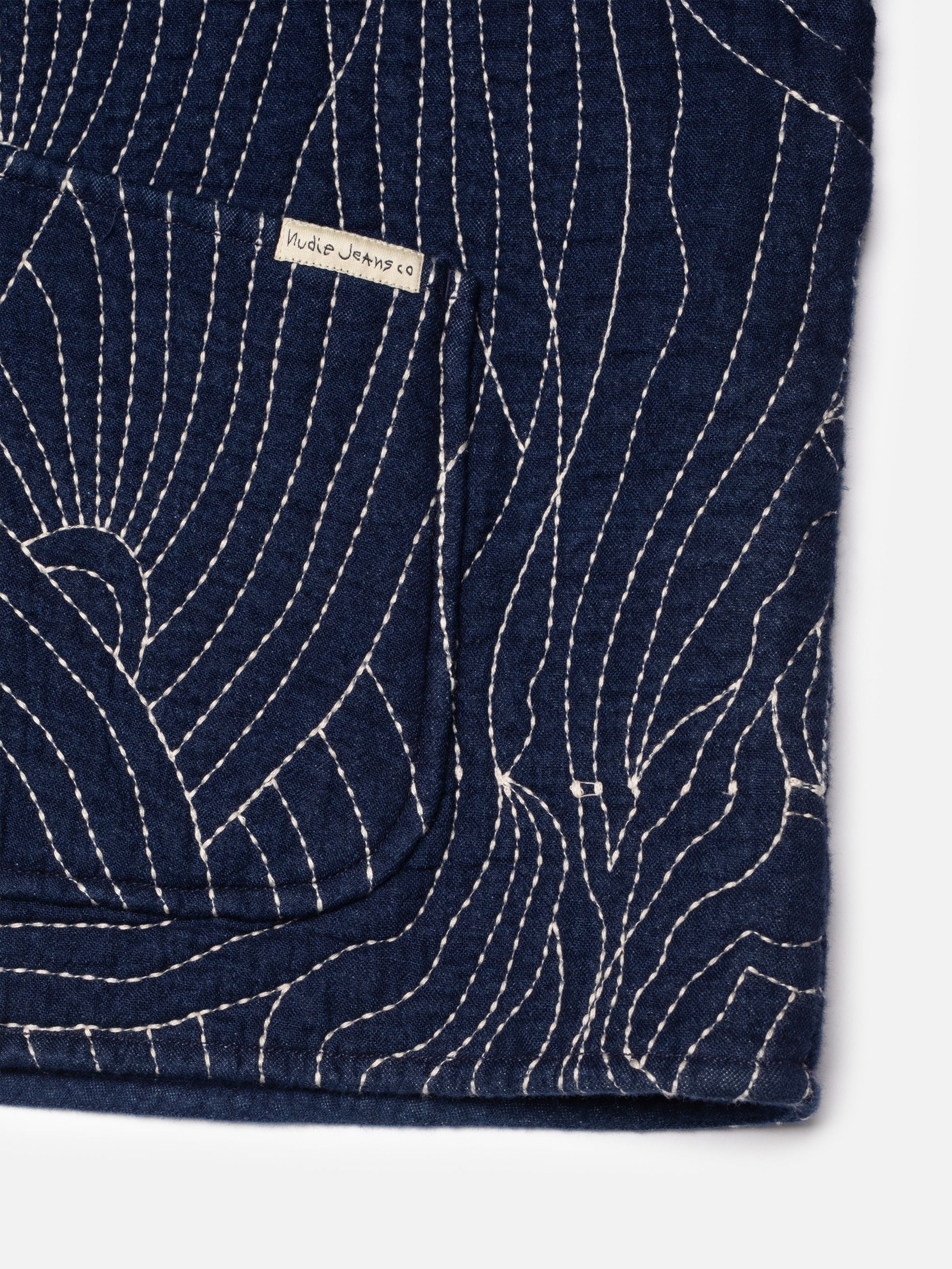 Nudie Jeans Lena Jacket Indigo Quilt, Nahaufnahme der gesteppten Indigo-Denim-Textur und der Vordertasche mit Logo-Etikett.