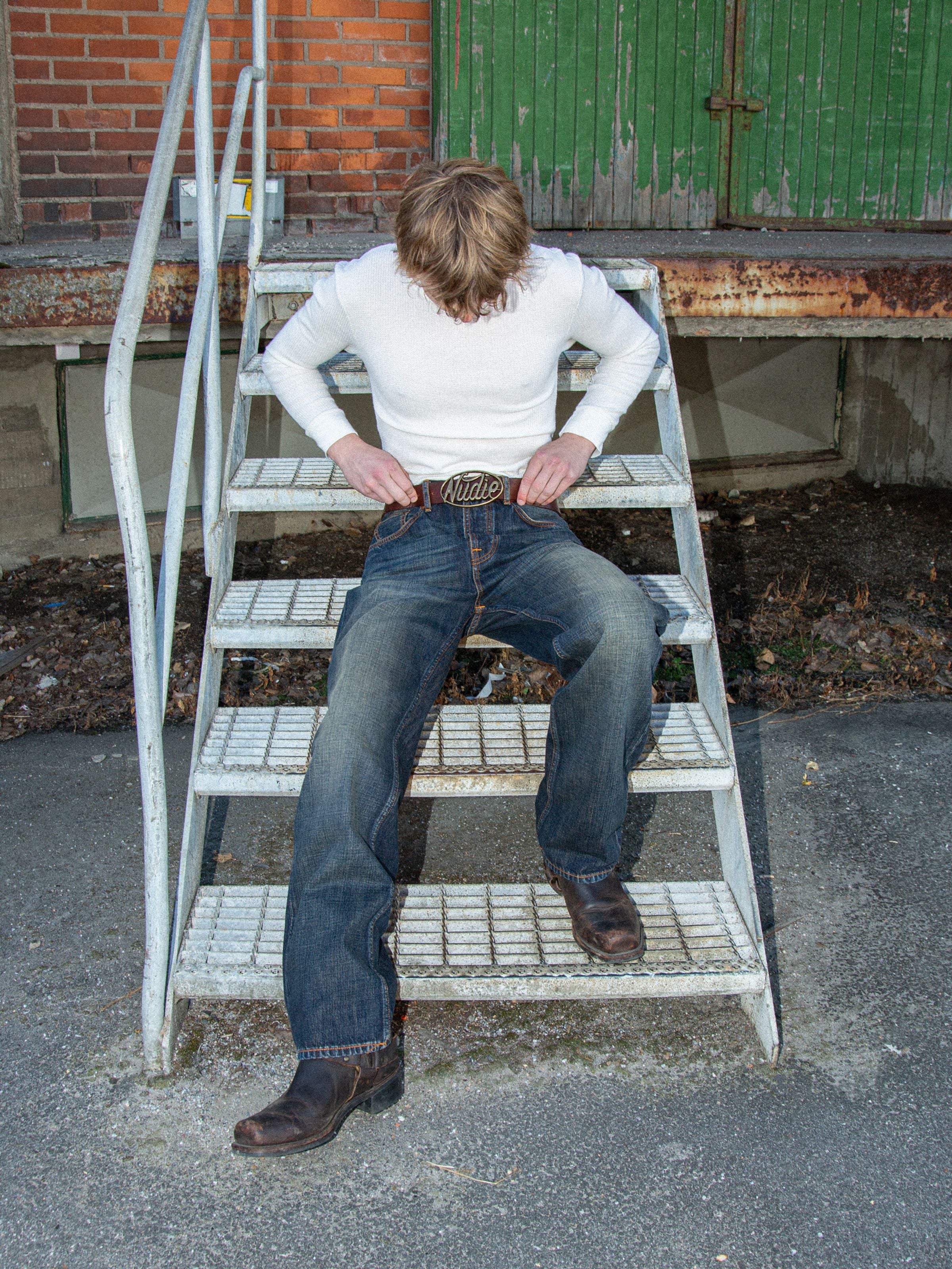 Nudie Jeans Tuff Tony Dirt Path, men's loose fit jeans shown in a lifestyle view sitting on metal stairs outdoors.