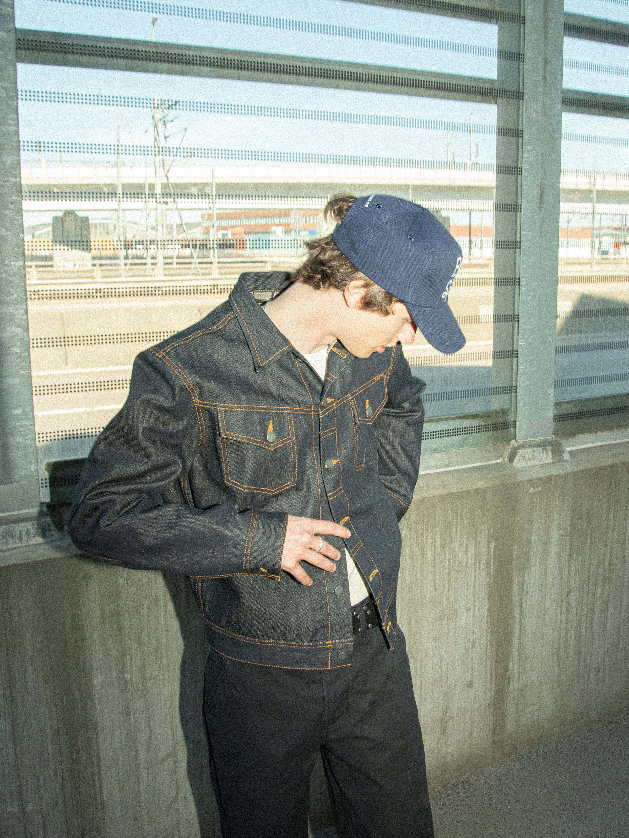Nudie Cap Bad At Sports in dark blue organic cotton, worn by a model with a denim jacket in an urban setting.