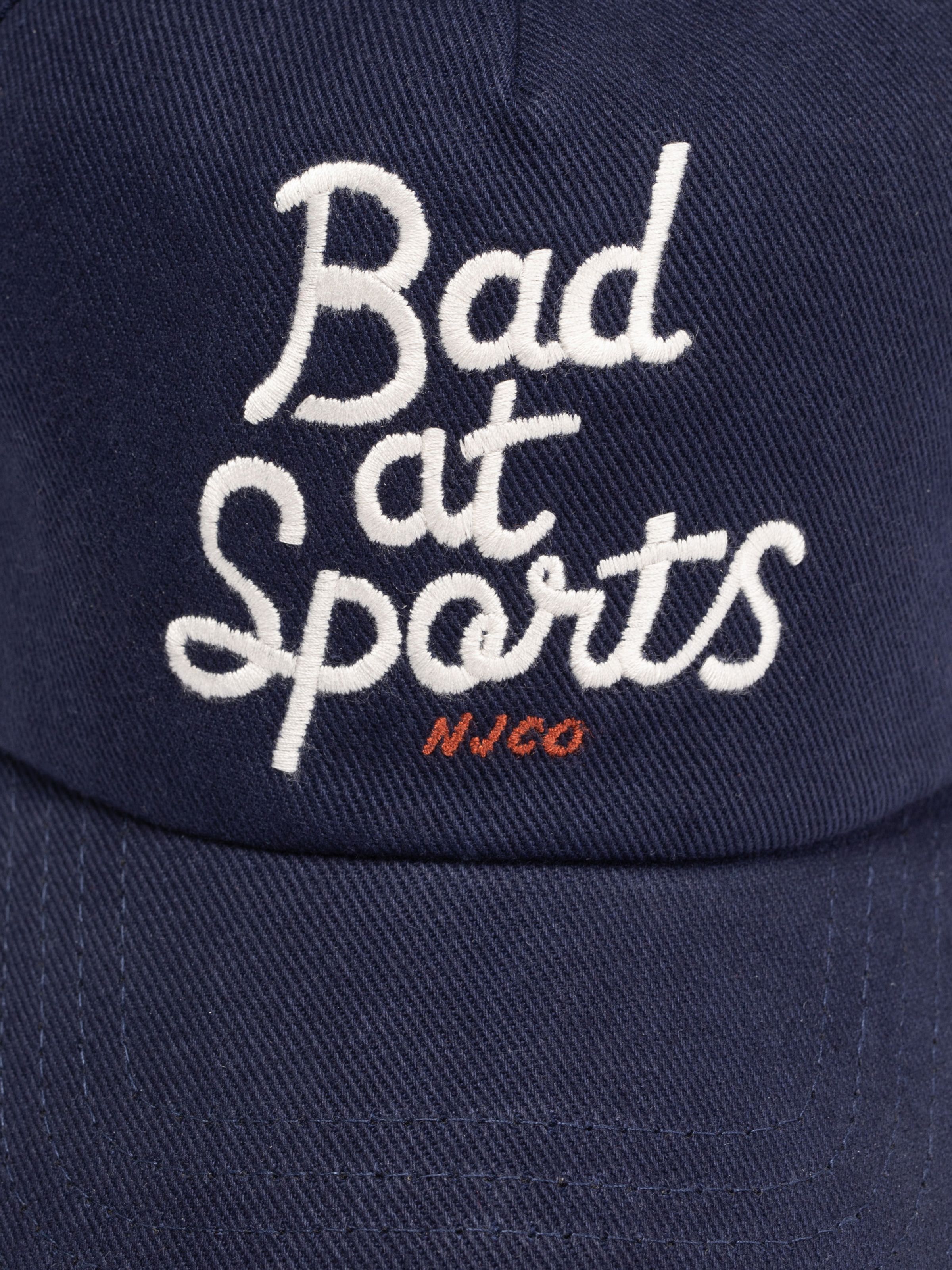 Nudie Cap Bad At Sports in blue organic cotton featuring white cursive script and orange NJCO embroidery.