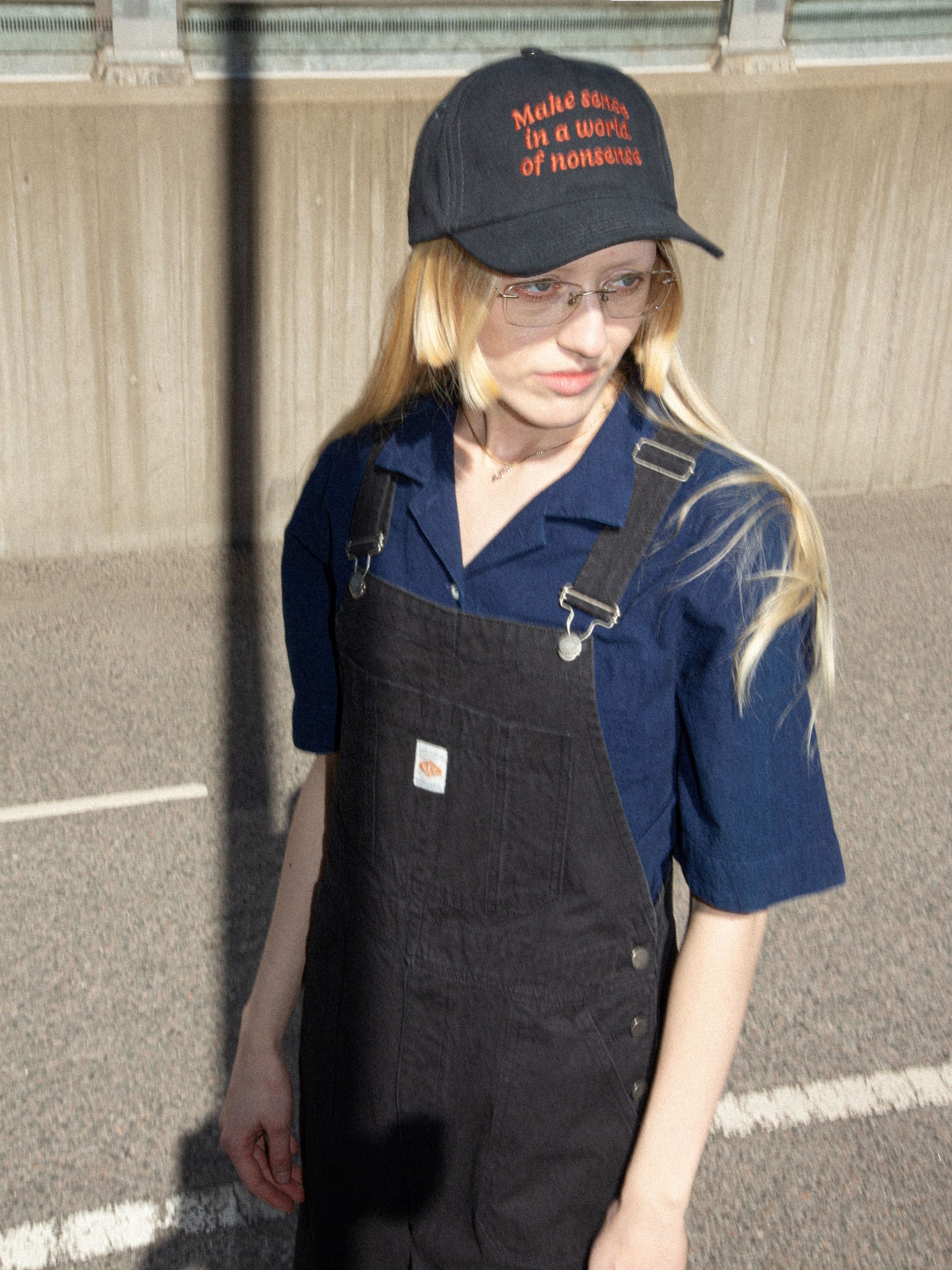Nudie Cap Make Sense in black organic cotton twill with red embroidery, worn by a model in black overalls.