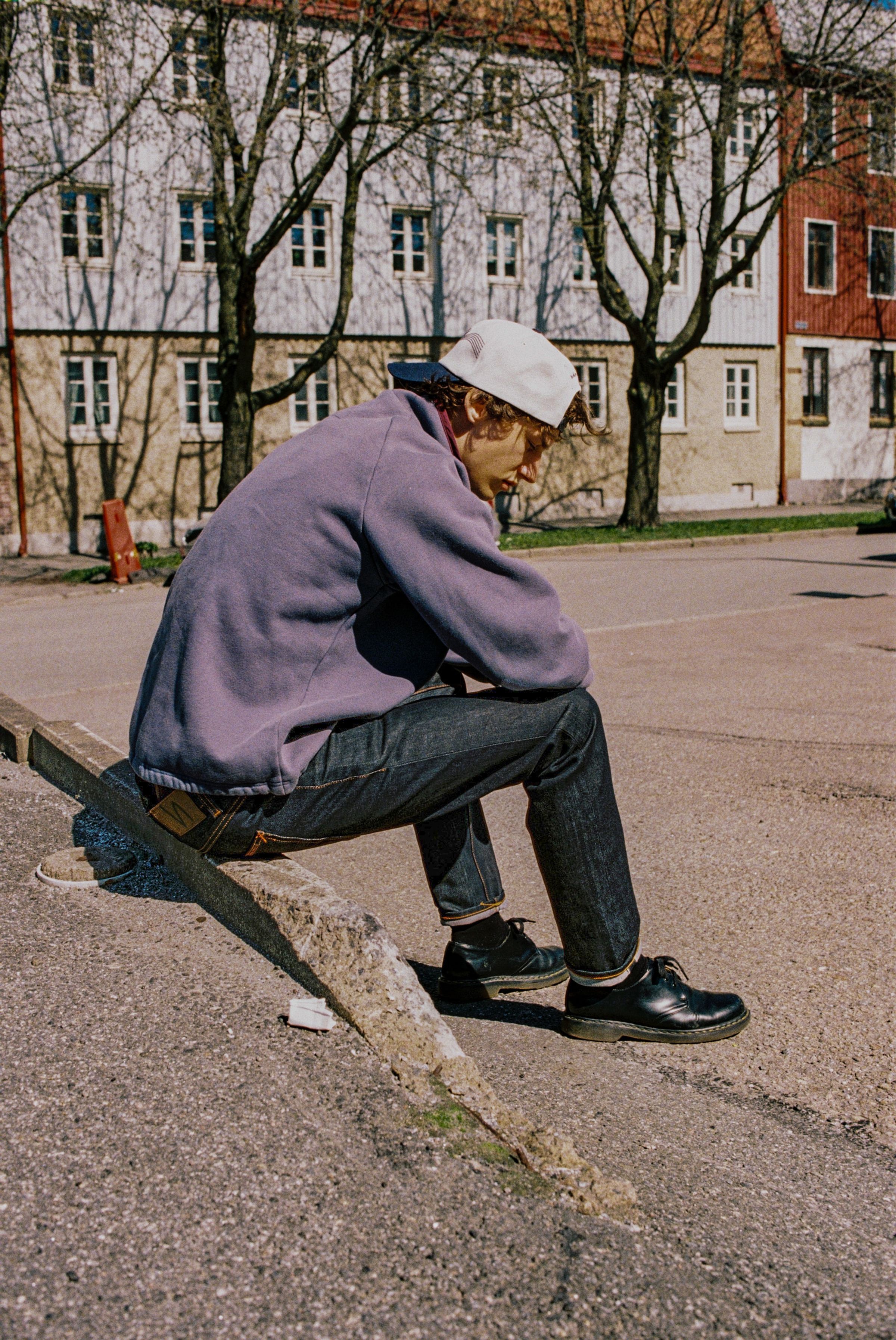 Nudie Cap Embo in Offwhite, a five-panel organic cotton cap worn backwards by a model sitting on a street curb.