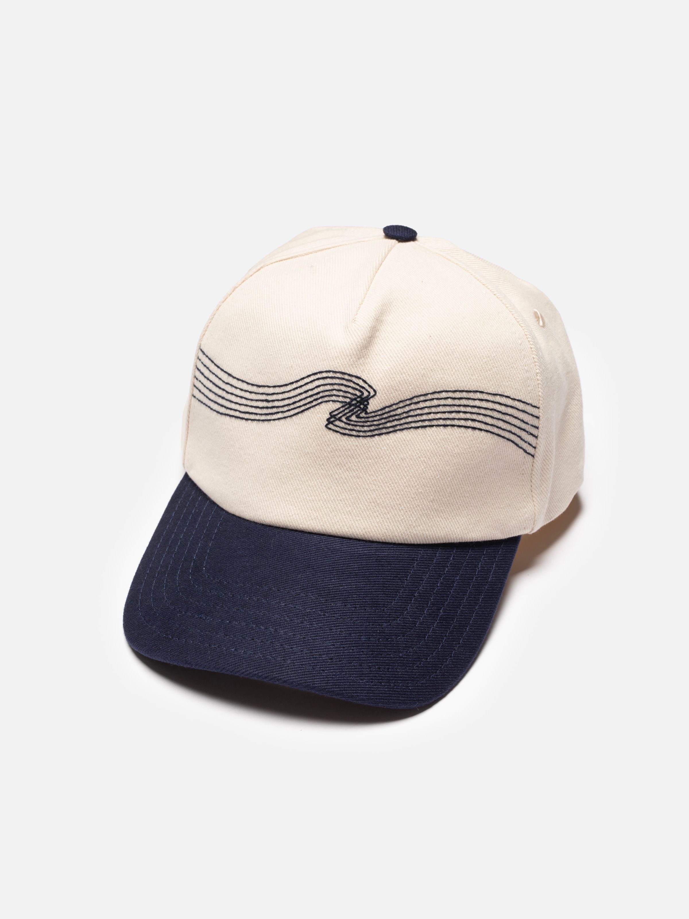 Nudie Cap Embo in off-white organic cotton twill, featuring navy wavy front embroidery and a dark contrast brim.