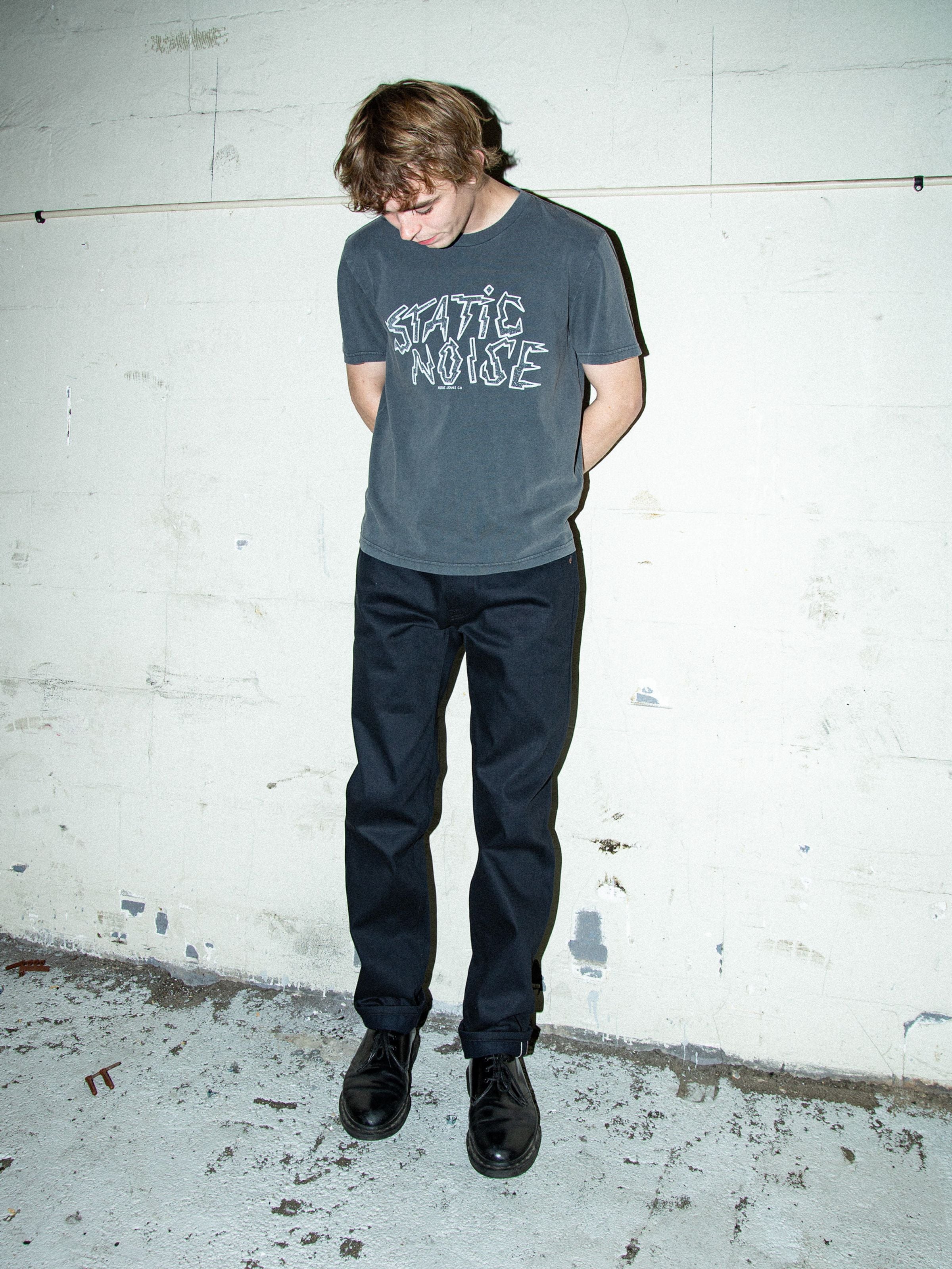 Nudie Jeans Gritty Jackson Dry Onyx Selvag, men's selvedge jeans in black, worn by a model standing against a textured wall.